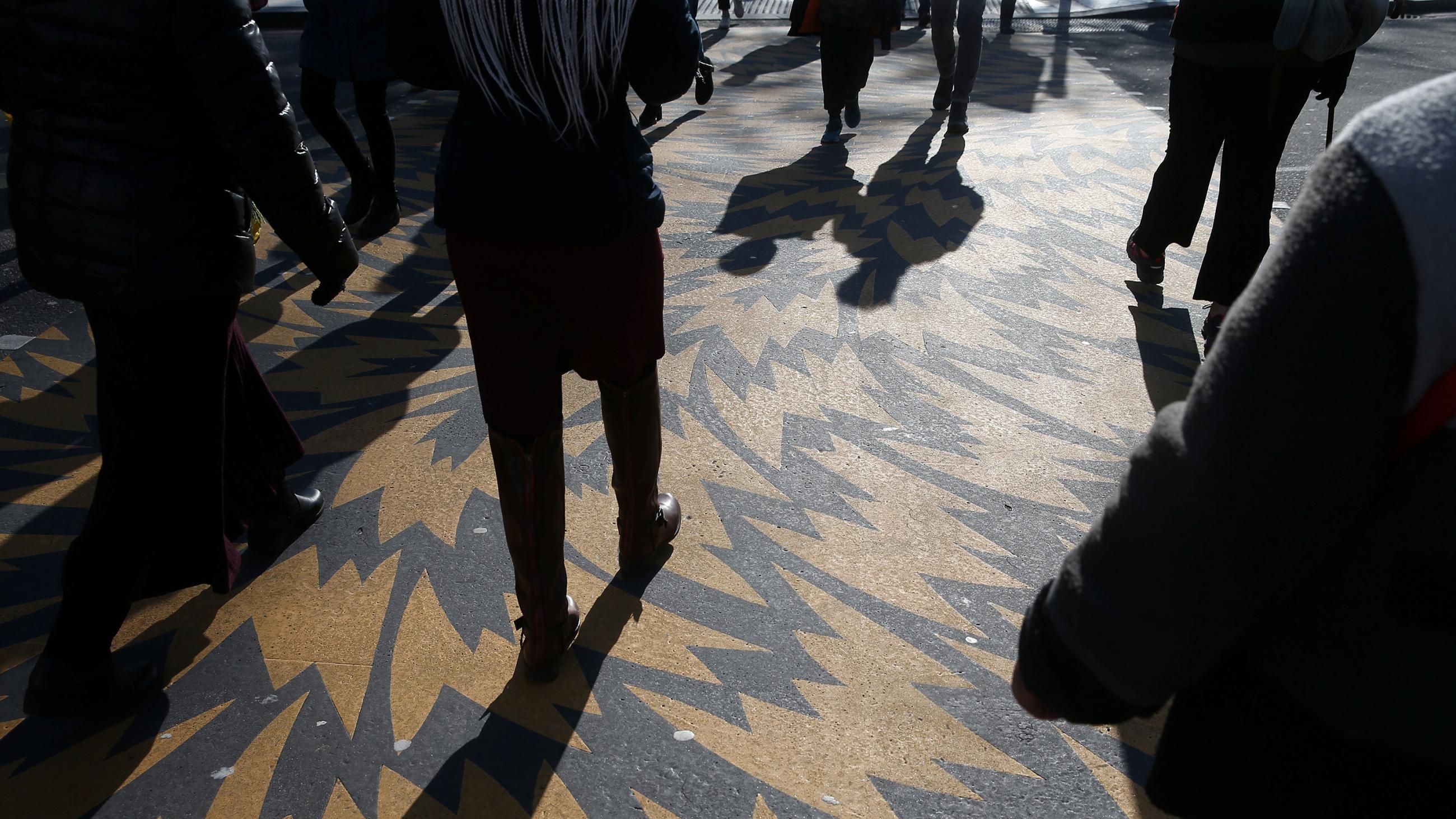 The photo shows the torso and legs of several people walking across a street over a decorative swirl pattern of paint on the crosswalk.