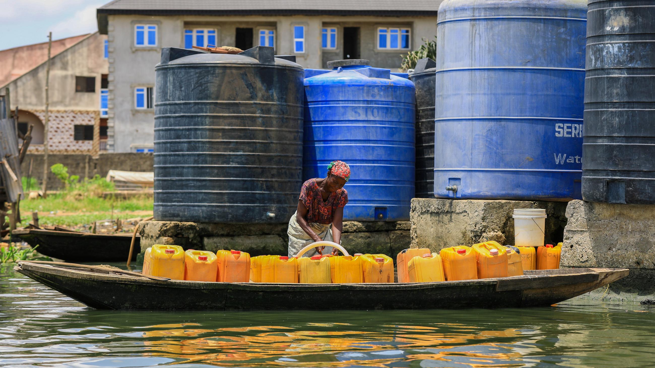 Picture shows a woman with a long dugout canoe pulled up to a filling station with a number of towering water tanks. She is filling a row of yellow jerrycans lined up inside the canoe. 