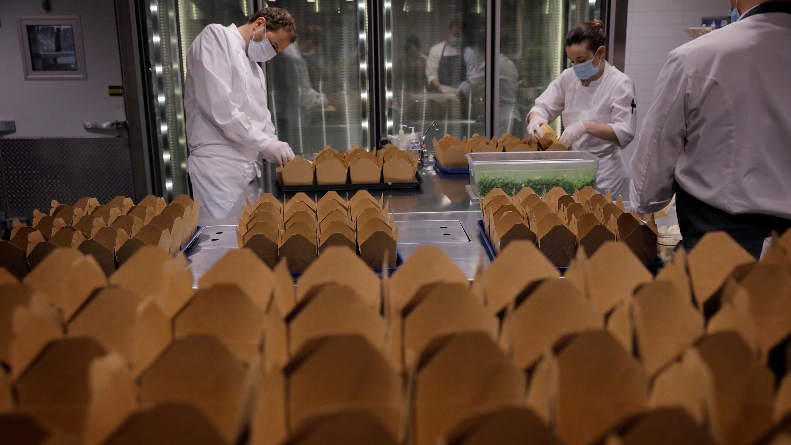 Picture shows a table filled with paper clamshell boxes open while chefs fill them with food.