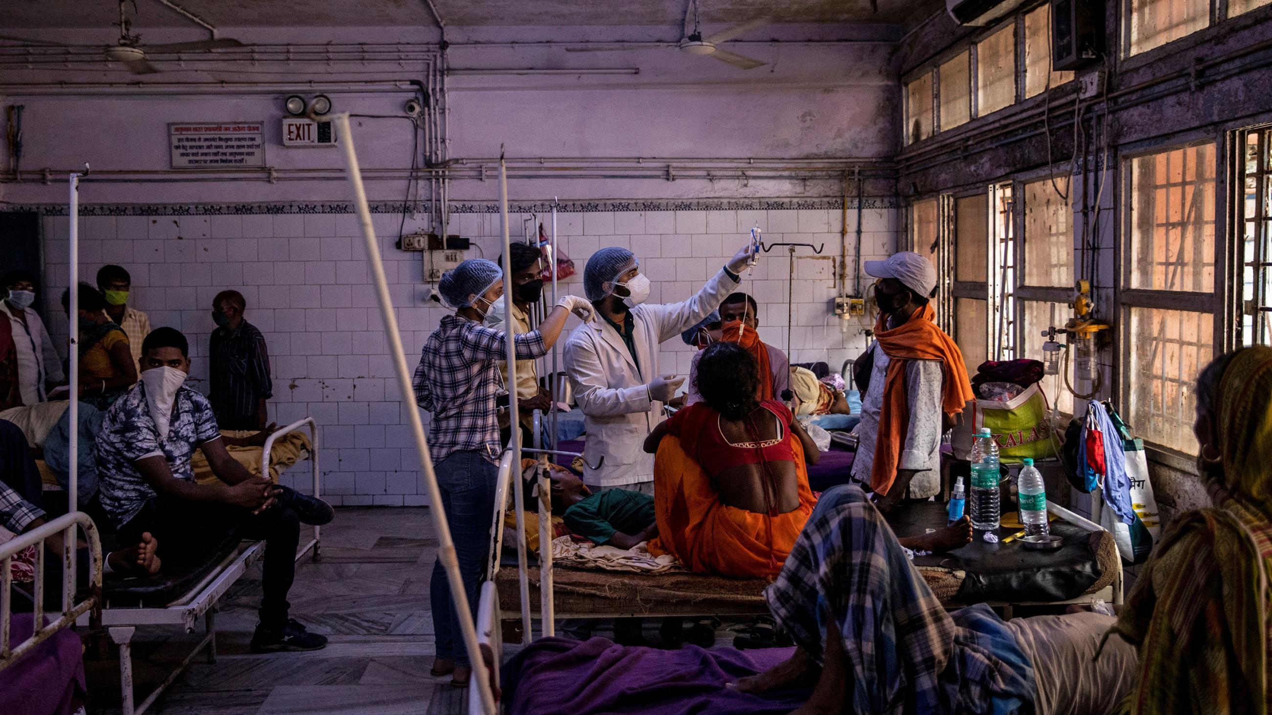 Photo shows a large ward bustling with people, including several seated in hospital beds. 