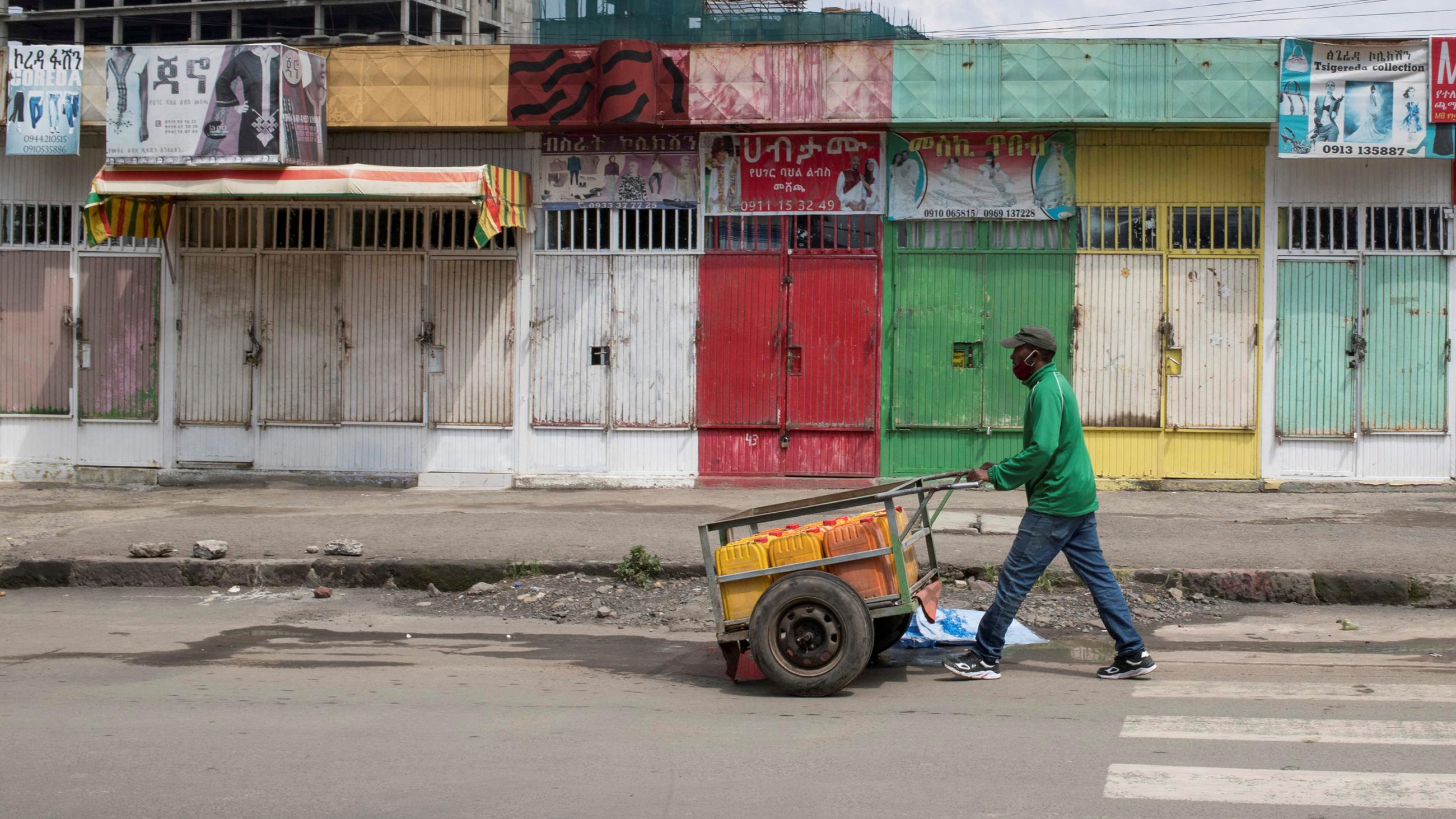 This is a striking photo of a man in a green shirt pushing a cart past wooden shuttered shops painted green and beige and red and yellow. 