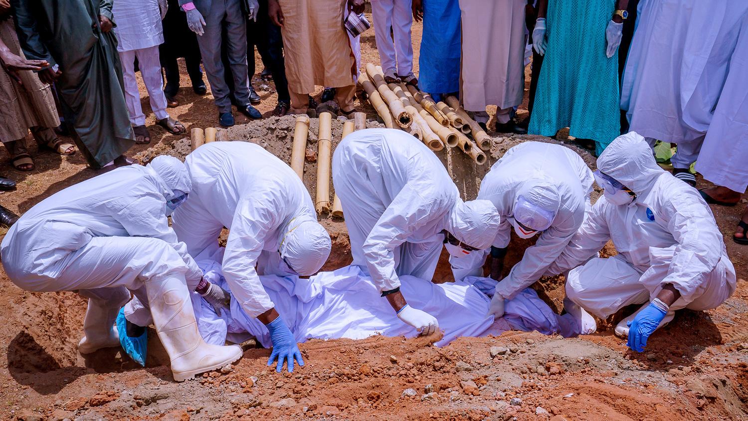 The photo shows people in protective gear lowering a body into the ground. 