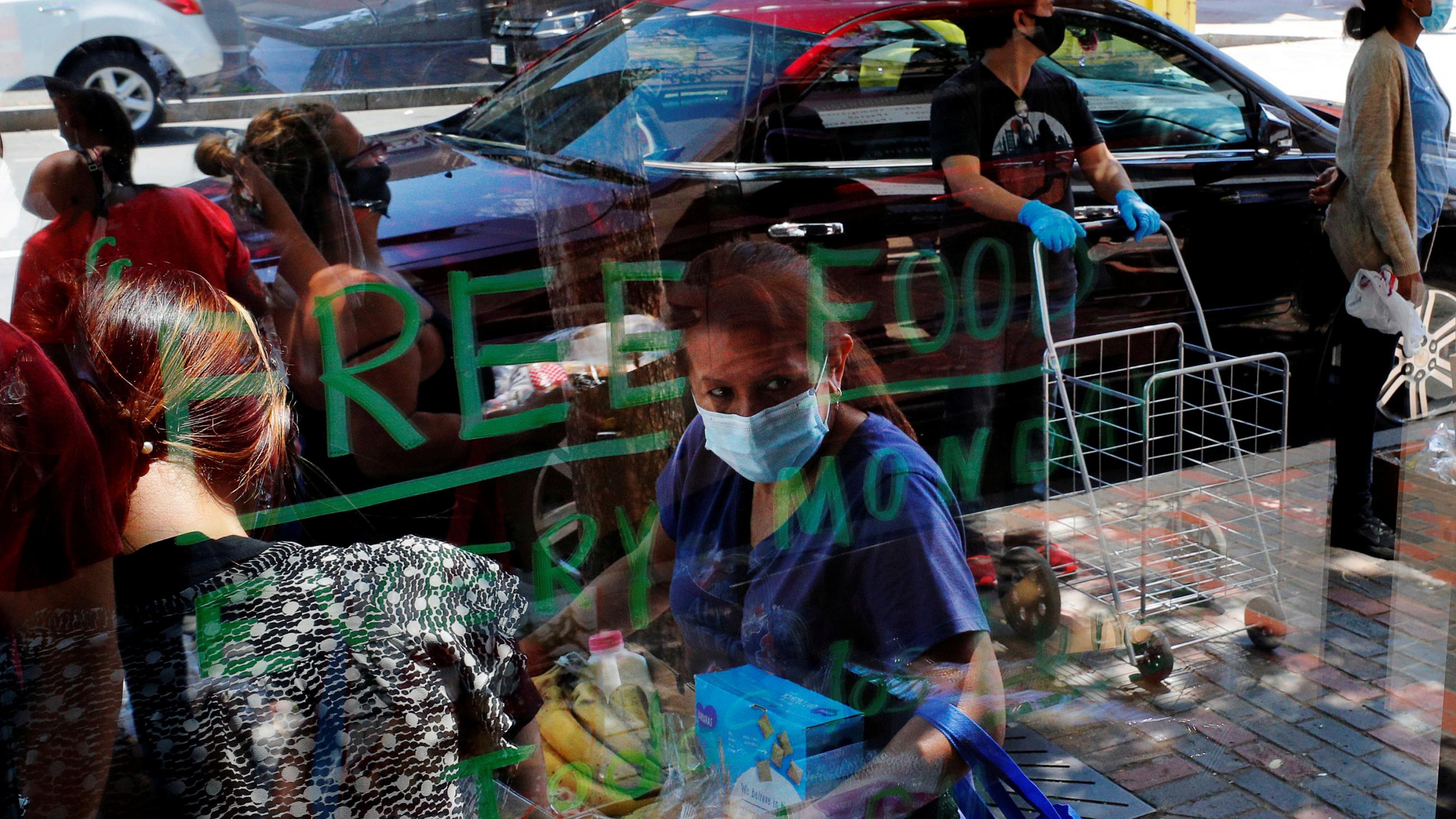 The photo shows people waiting in line while others leave with armloads of food from the other side of a plate glass storefront on which is scrawled in hand-written green letters "Free Food Every Monday." 