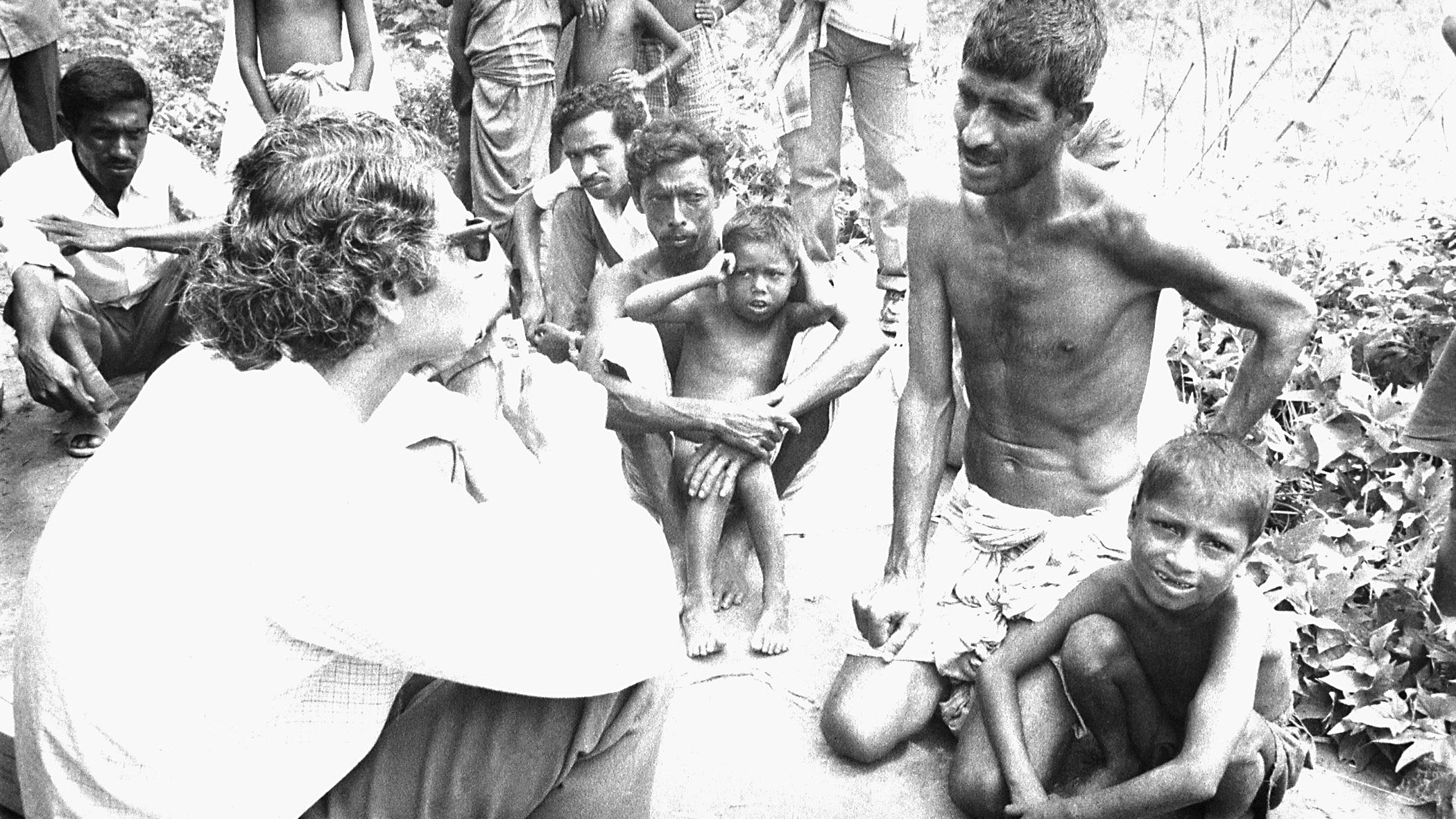 The photo shows the doctor seated on the ground speaking with a number of adults with several children also seated amongst them.