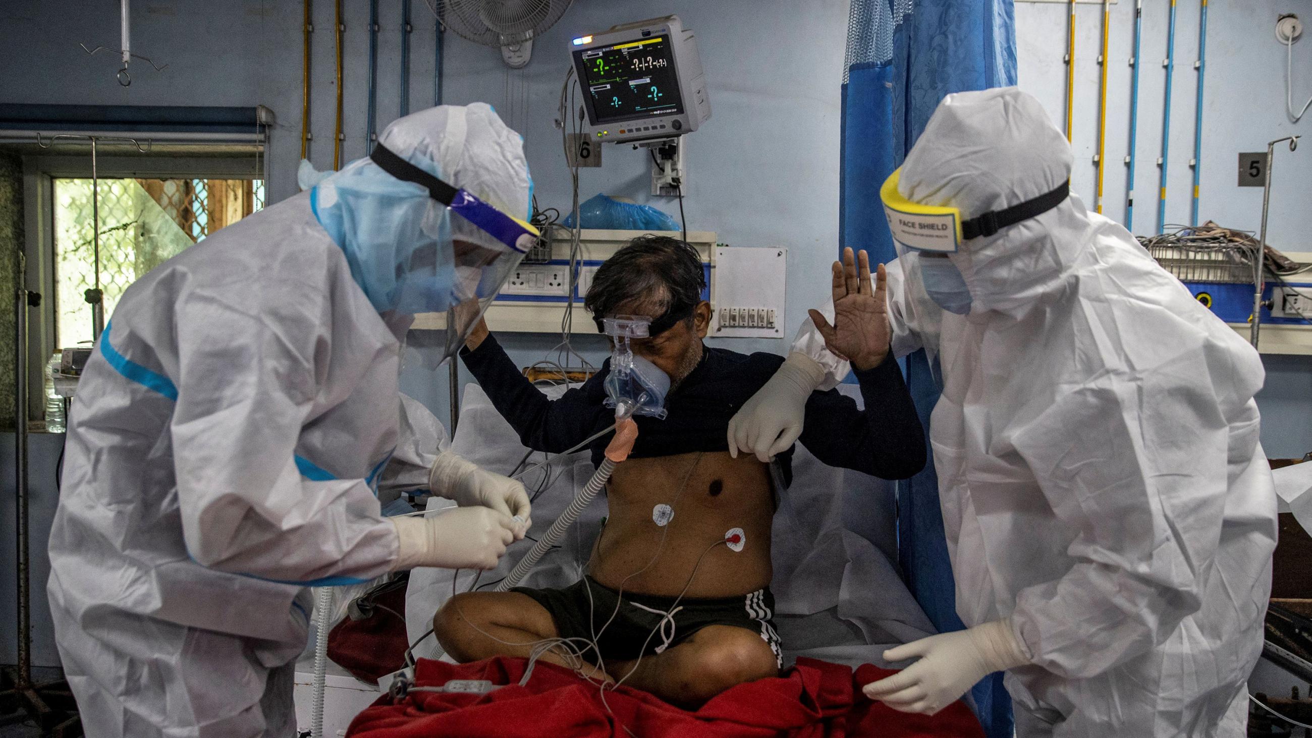 The photo shows two workers wearing protective gear tending to a patient sitting cross-legged on a hospital bed. 