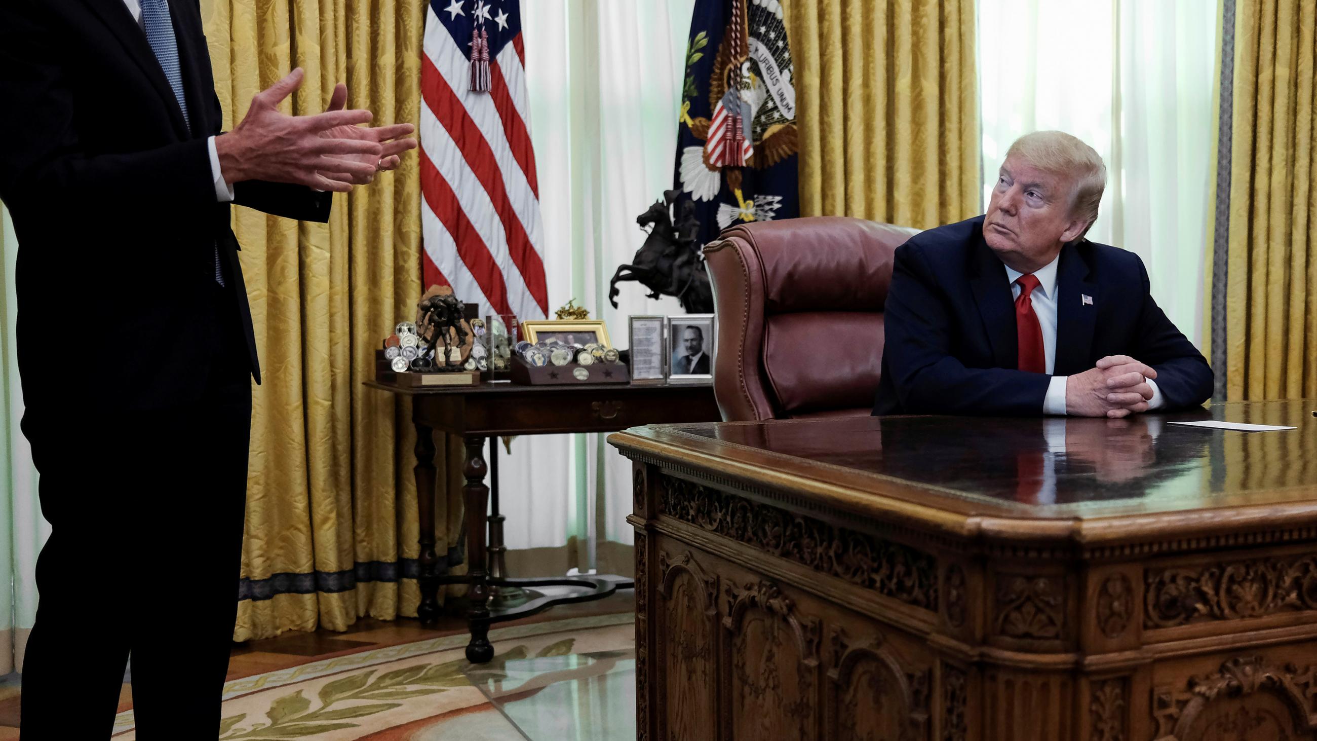 The photo shows the president at his desk looking to the right as an suited man speaks.