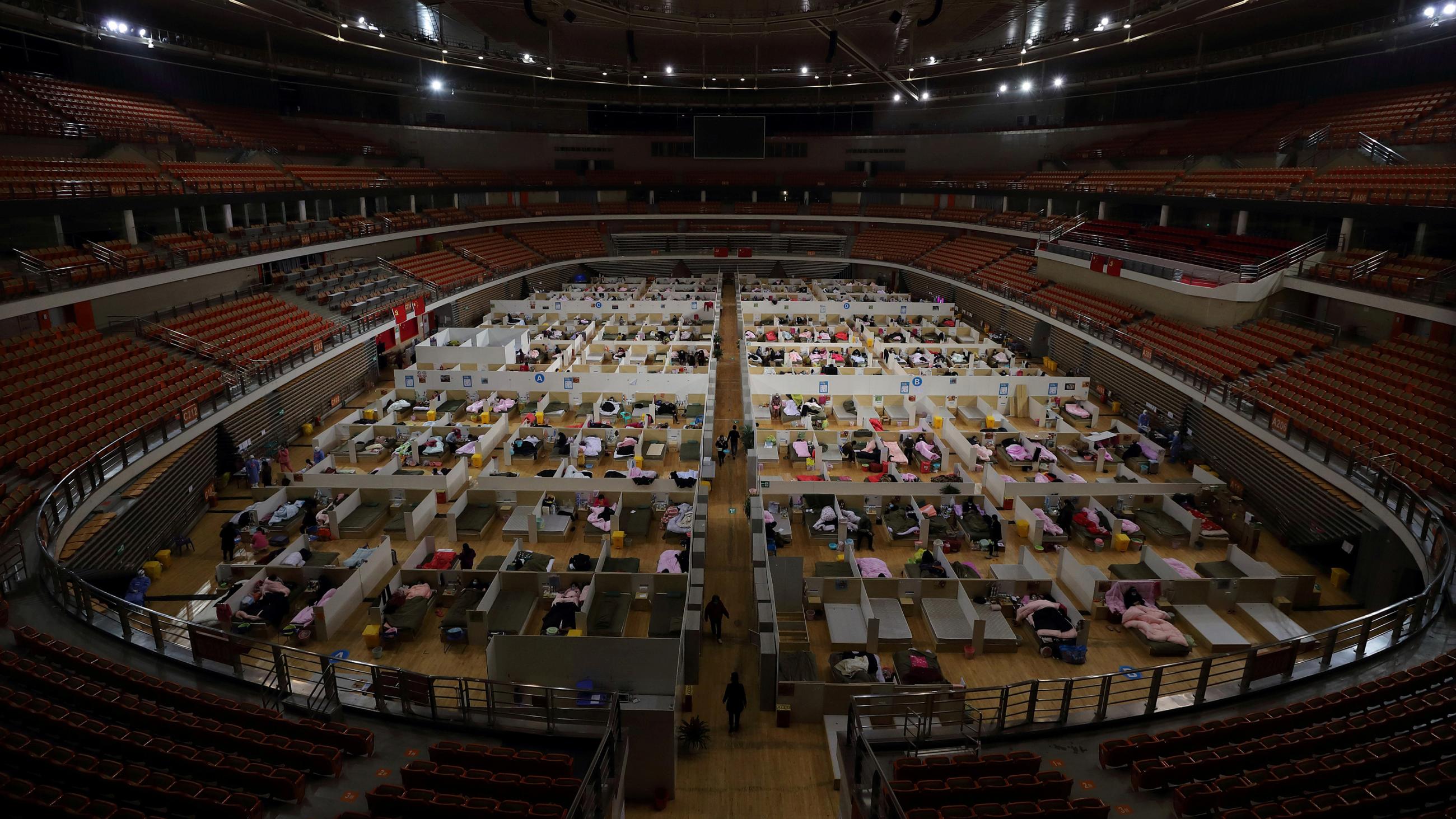 Picture shows a stadium with empty seats and a floor packed with patients resting on mats. 