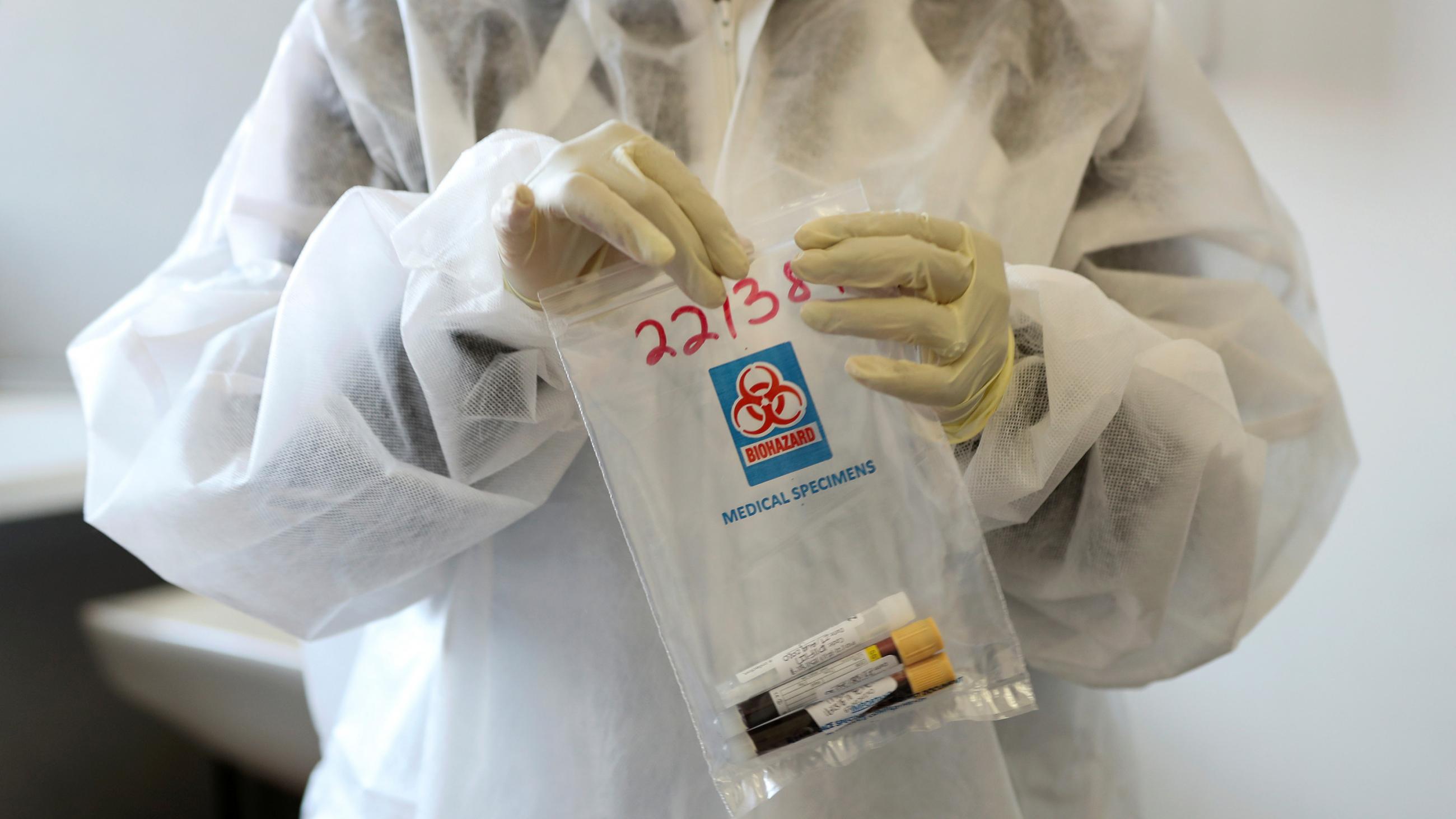Picture shows a health worker wearing protective gear holding a clear plastic bag with vials of what appear to be blood. 