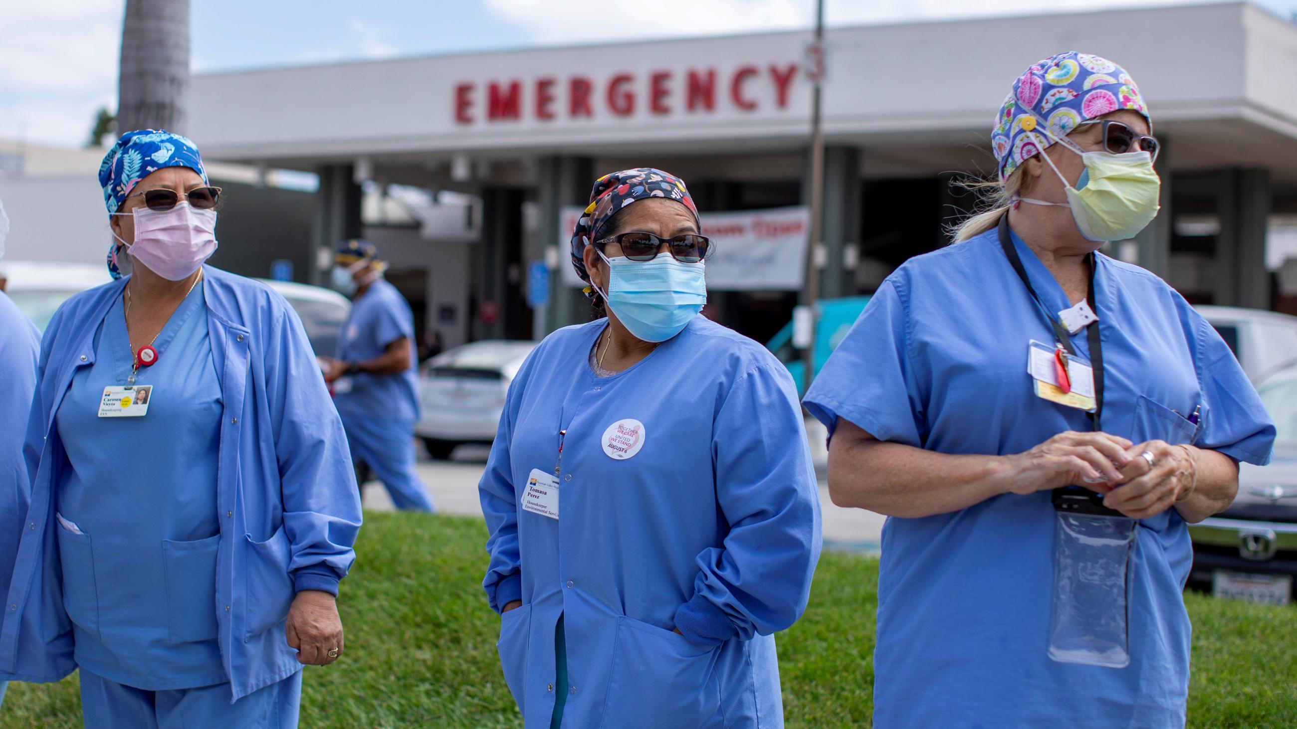 The photo shows a collection of health workers in blue scrubs protesting in front of a hospital entrance adorned with the word "EMERGENCY." 