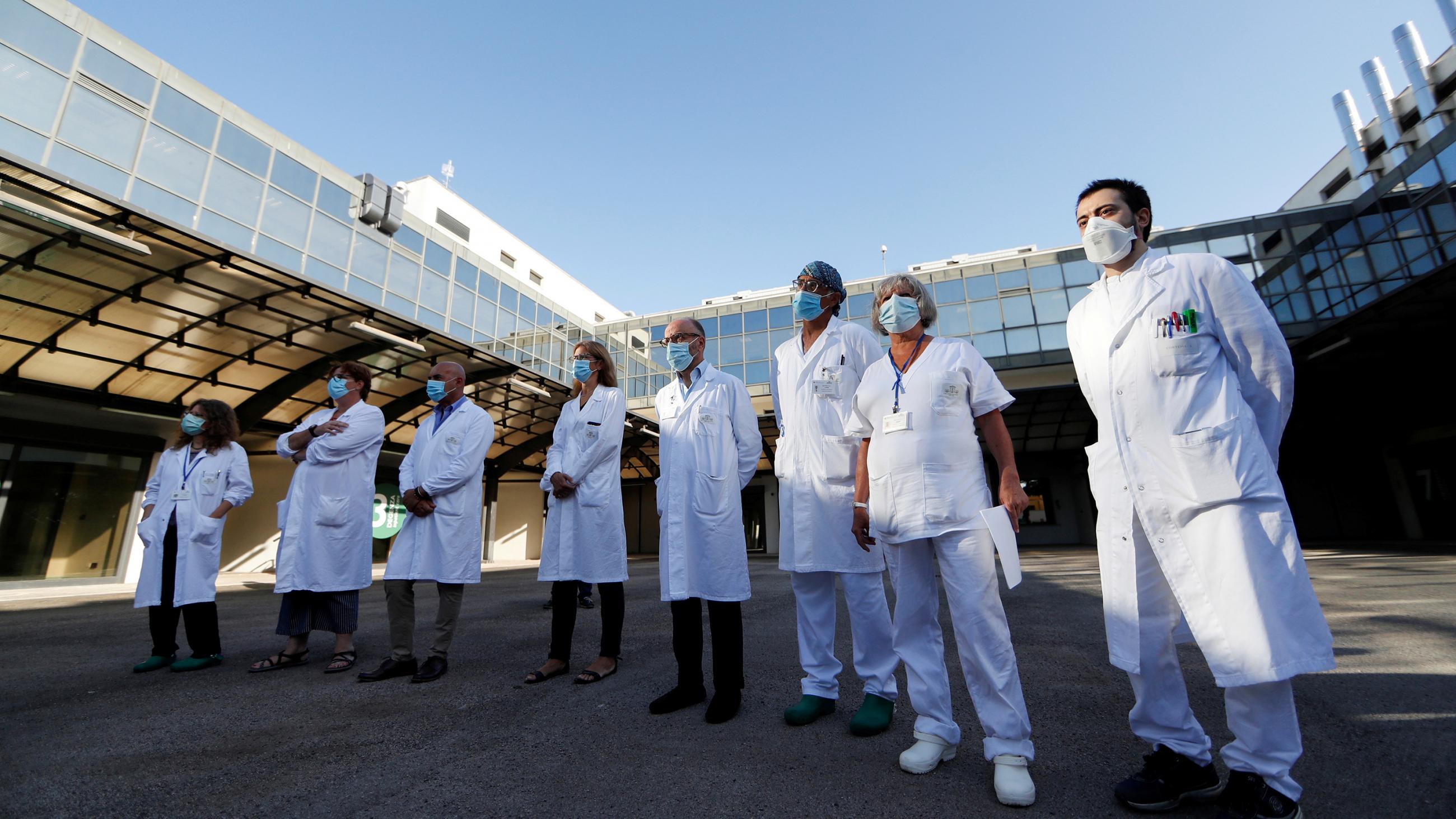 The photo shows a line of medica workers with masks and white coats in front of a modern hospital on a clear day. 