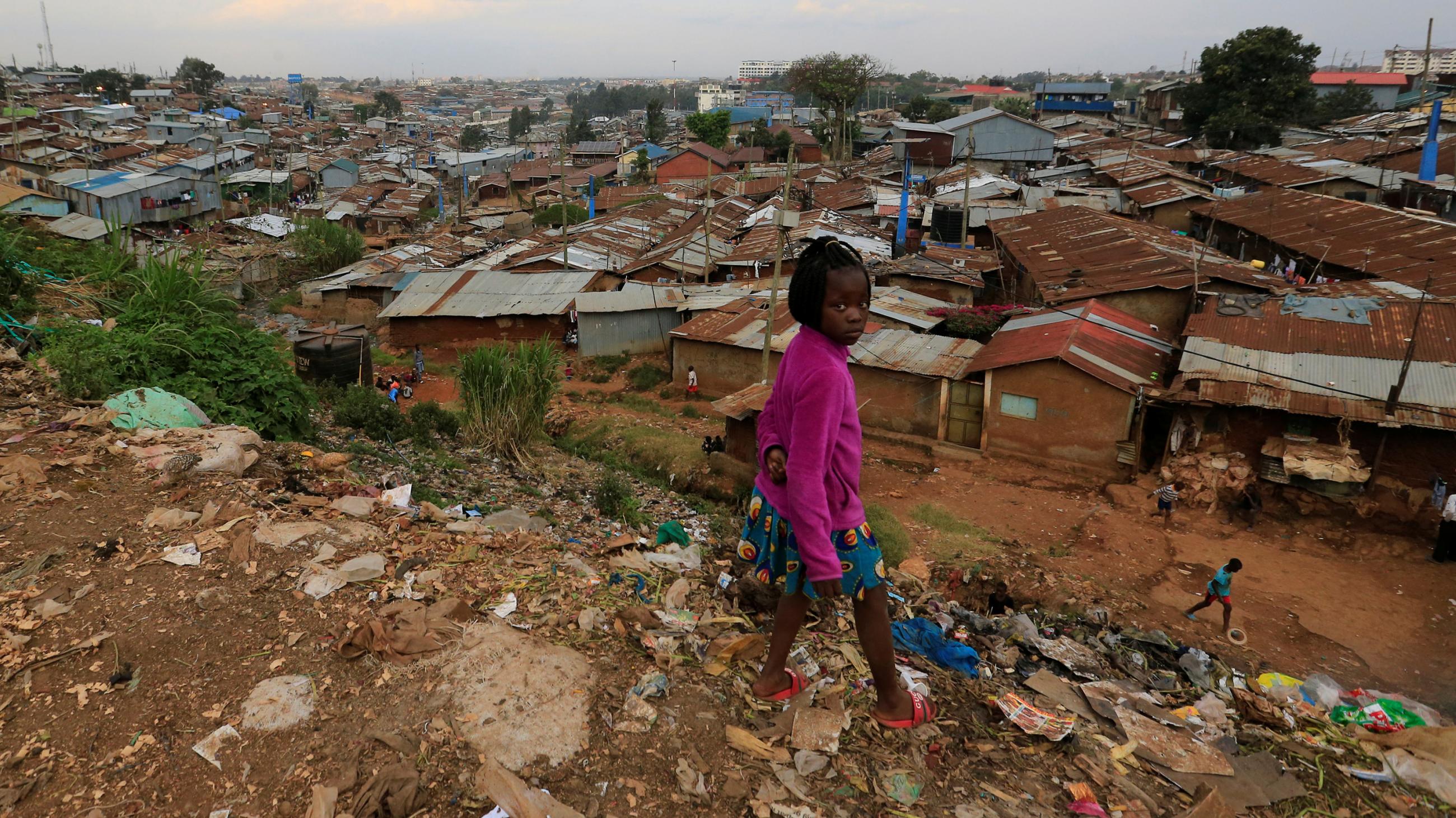The photo shows a girl standing atop a hill littered with trash overlooking a residential area of poor houses with rusty metal roofing. 