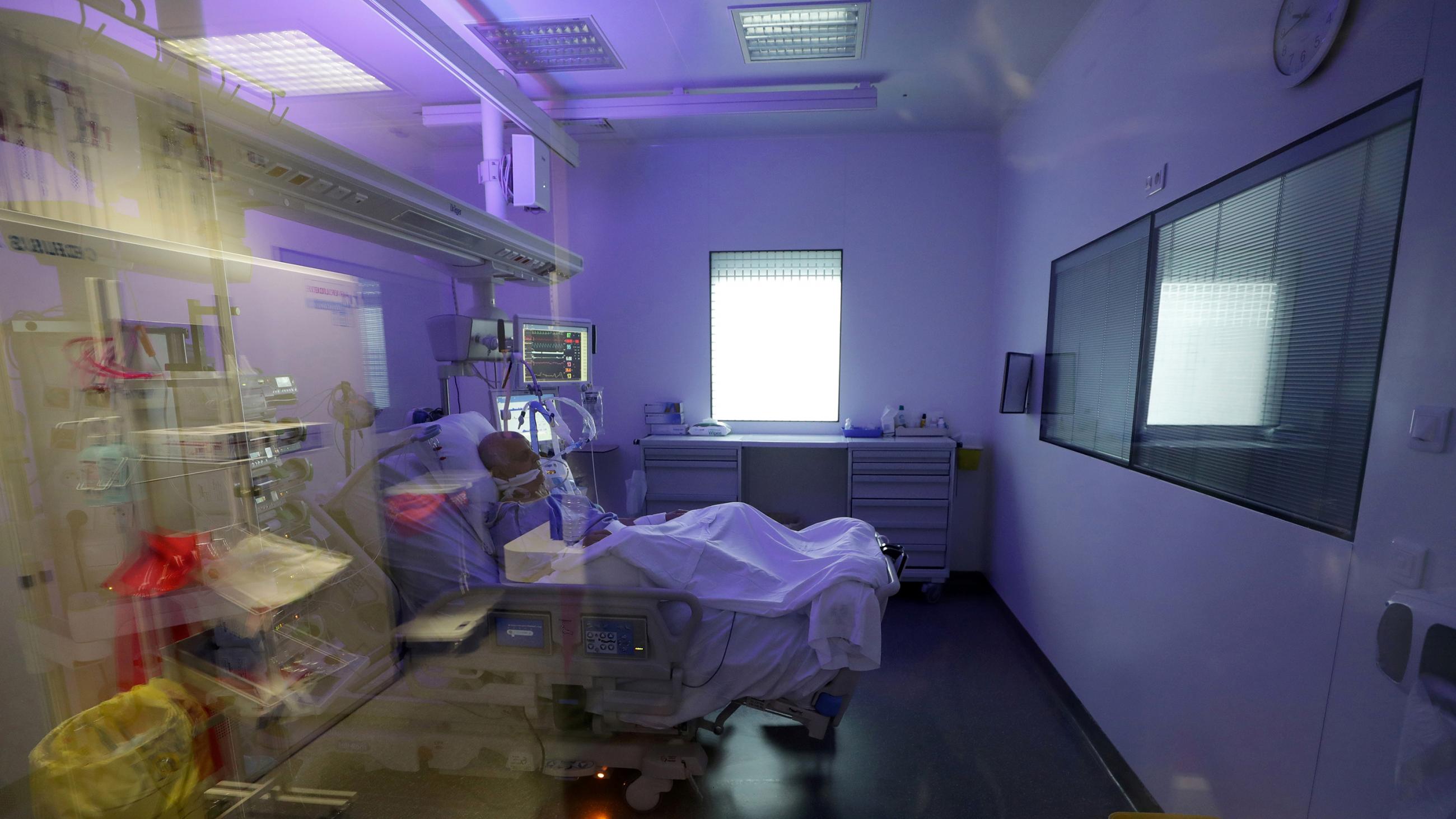 The photo shows a hospital room with a patient in a bed in the middle. The room is bathed in purple light.