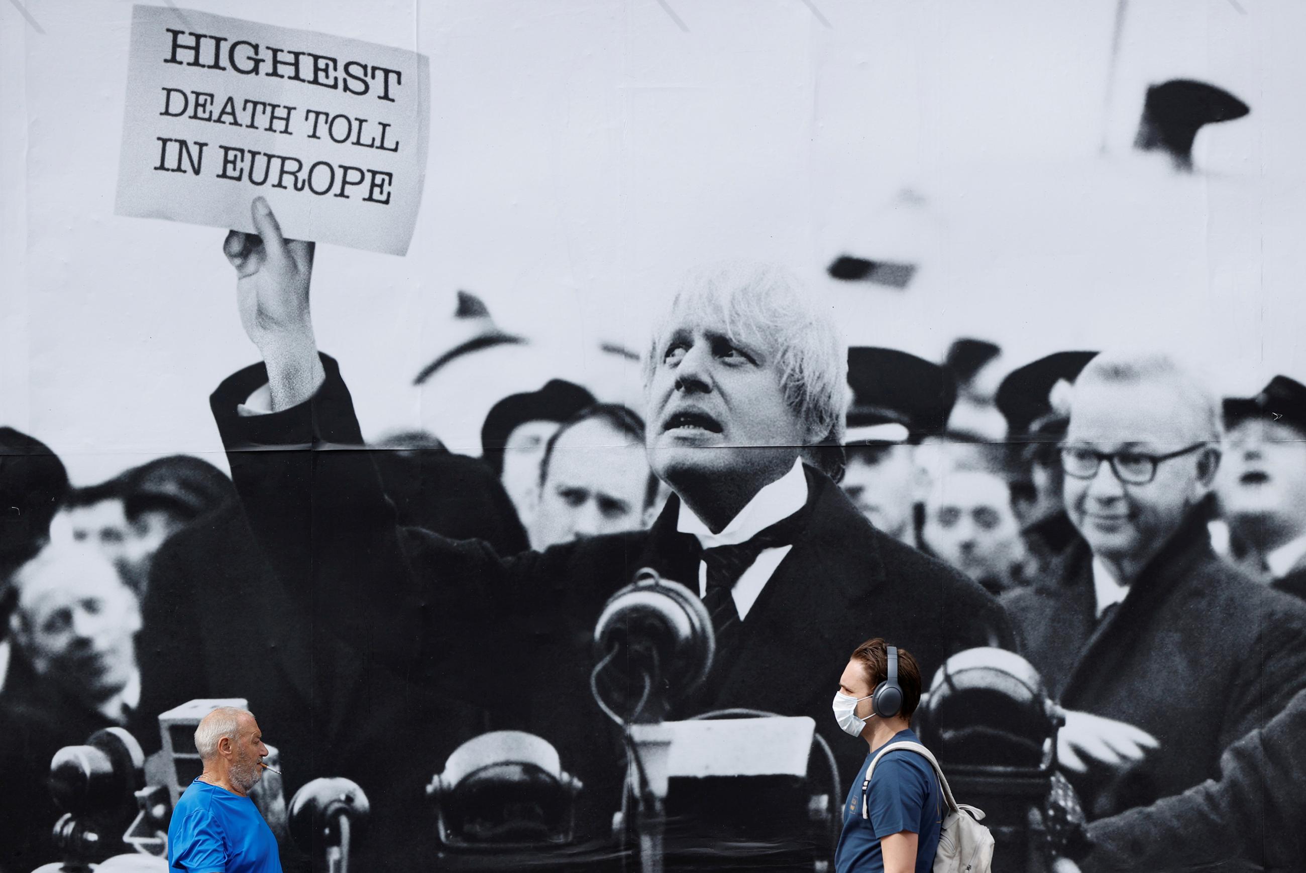 The photo shows a few people walking by a huge sign with a black and white picture of the PM holding up a placard that reads, "Highest Death Toll in Europe." 