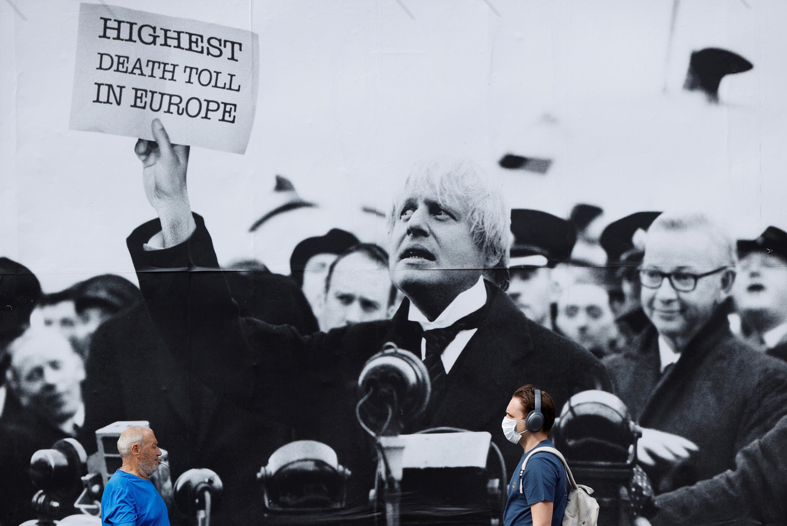 The photo shows a few people walking by a huge sign with a black and white picture of the PM holding up a placard that reads, "Highest Death Toll in Europe."