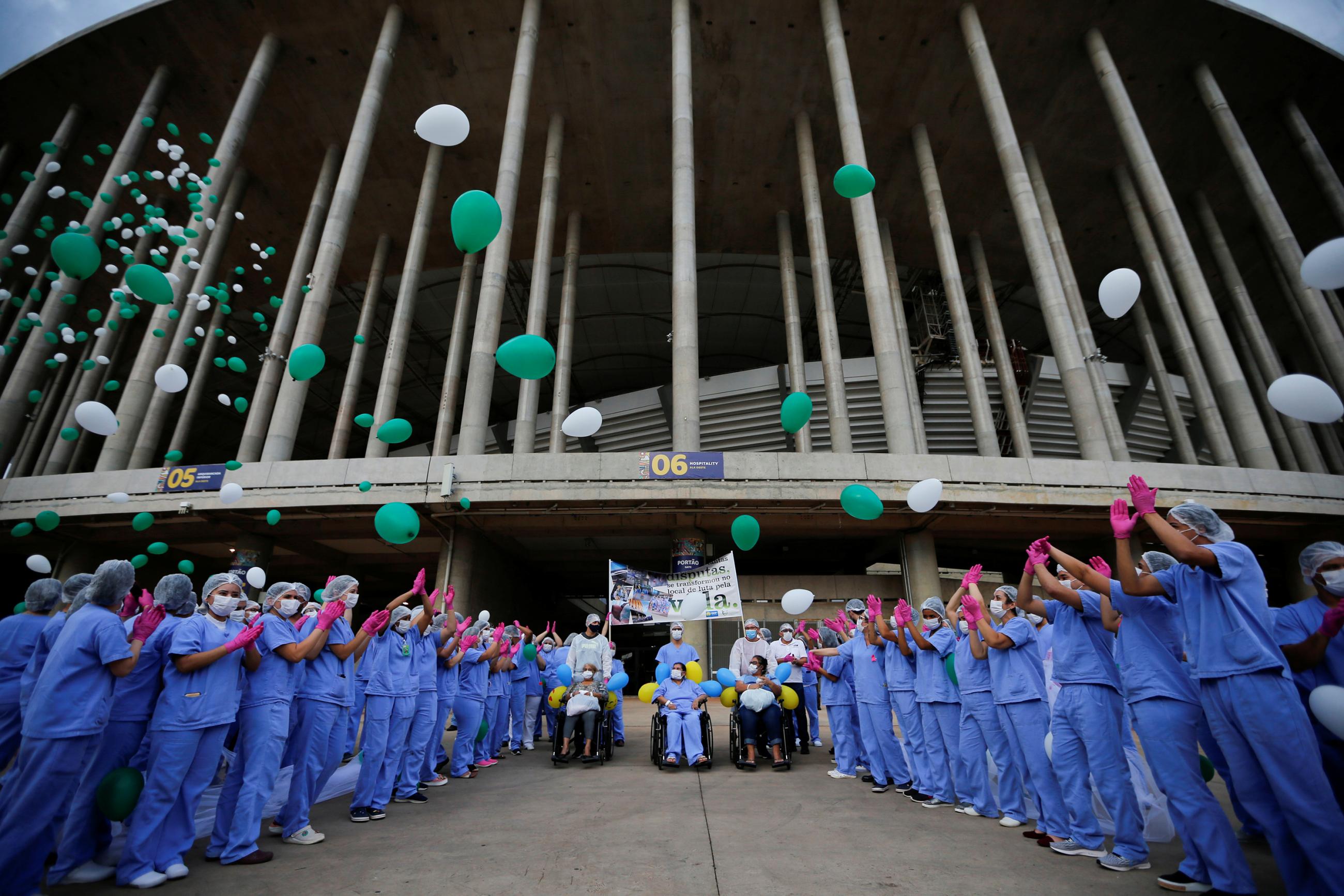 The photo shows a large crowd of blue scrubs wearing health workers cheering and releasing green and white balloons at the entrance of a large stadium. 