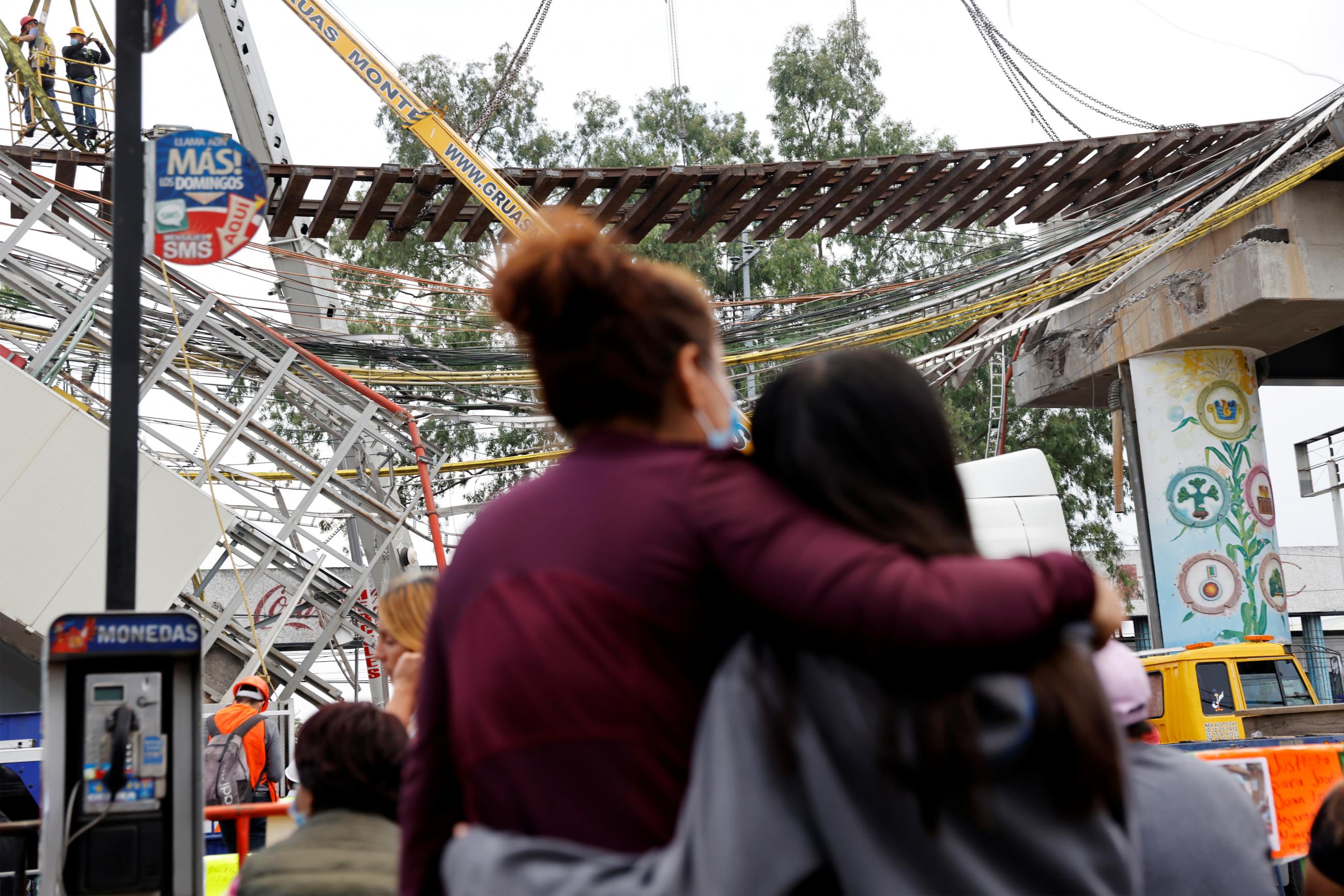 People look at employees from different companies working at the site where an overpass for a metro partially collapsed with train cars on it, in Mexico City, Mexico, on May 11, 2021. 