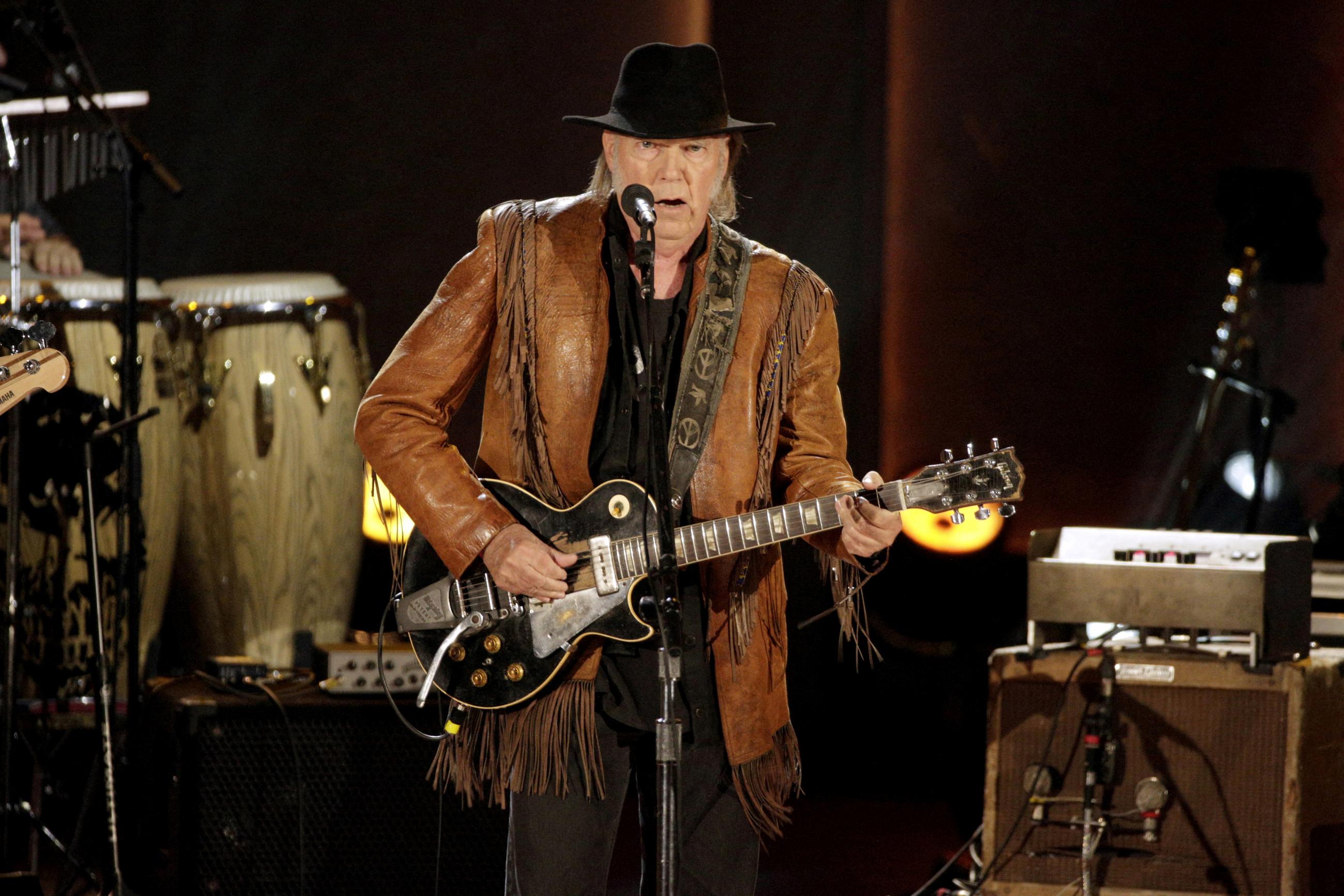 Singer-songwriter Neil Young plays guitar on a stage wearing a fringed jacket and black hat.