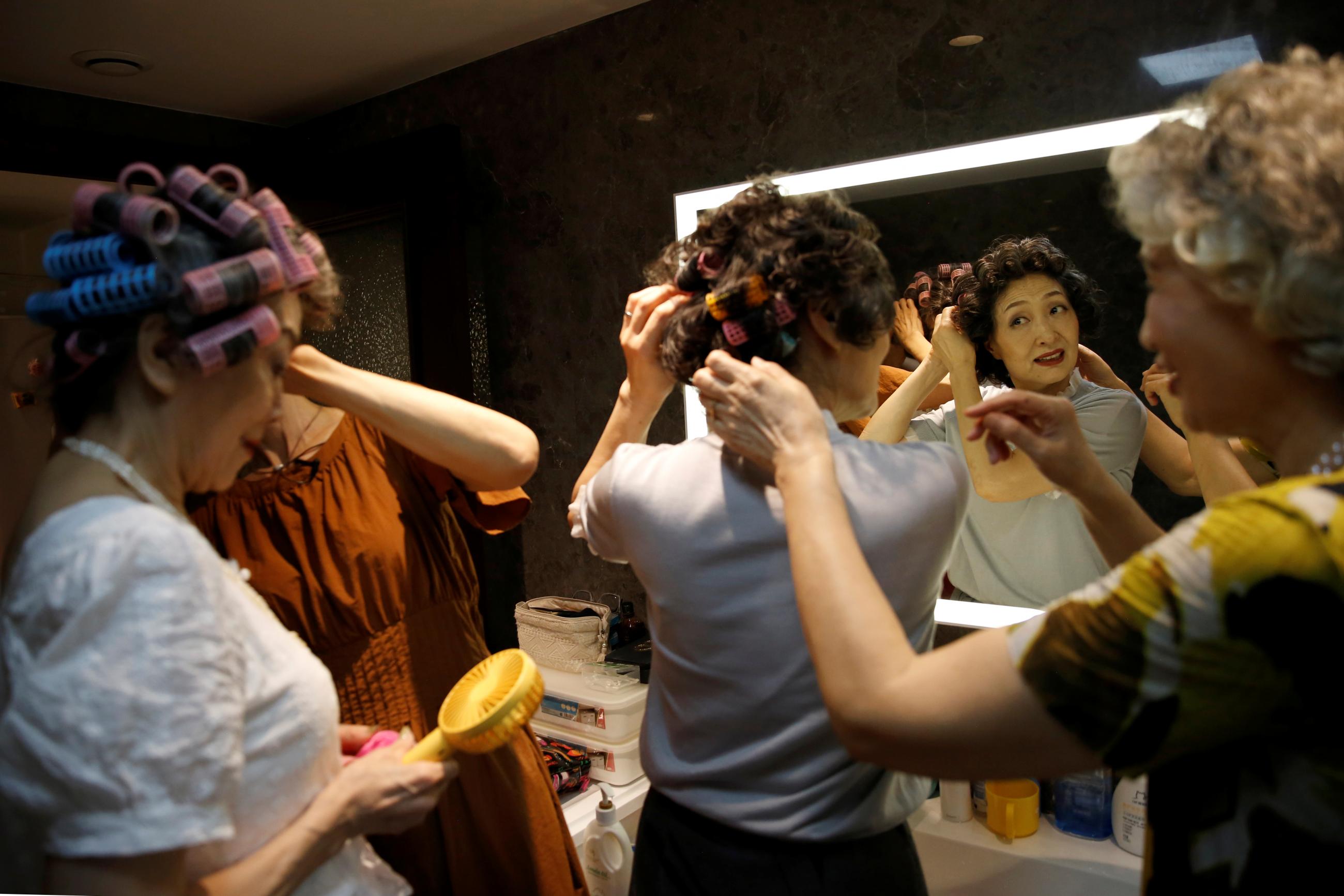 A group of older ladies prepare their hair in a mirror in preparation for a video shooting