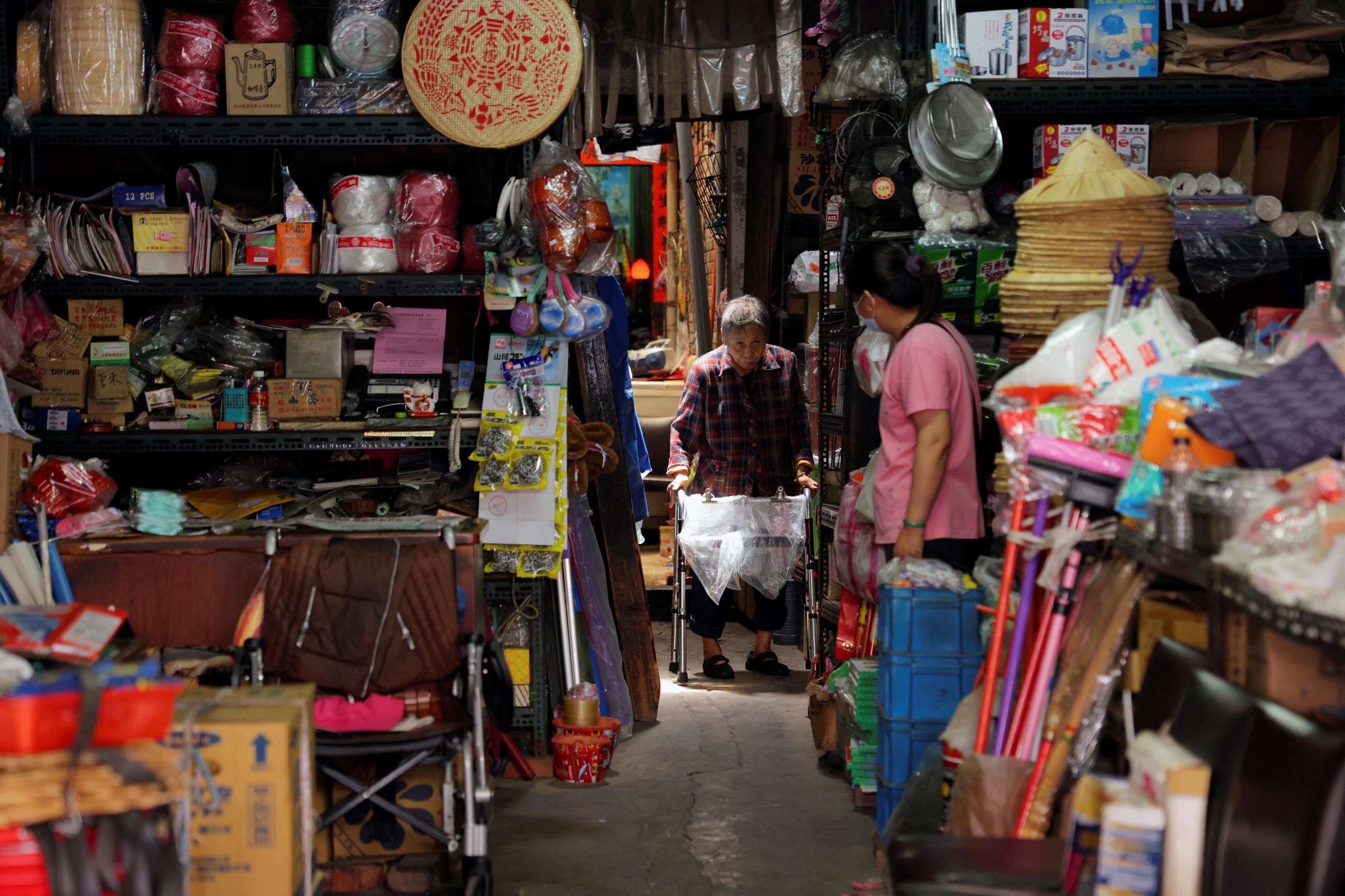 An elderly woman walks with a walker through a doorframe in the back of a hardware store, on either side of which are shelves laden with goods in New Taipei, Taiwan on April 22, 2022. 