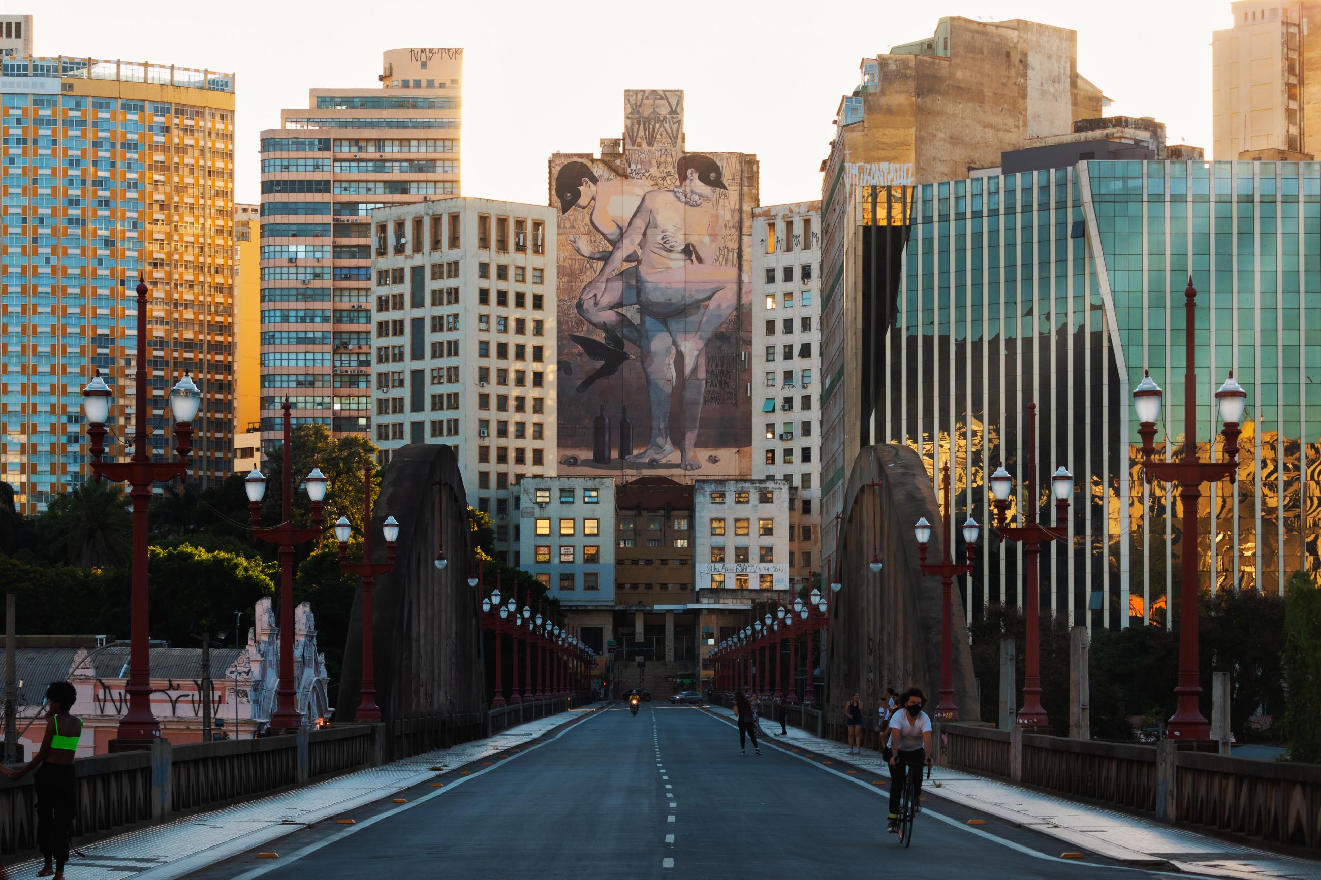 People walk below the arches of the Viaduto Santa Tereza bridge, adorned with red lamp posts, which frame a mural of two people wearing black bird masks adorning a cityscape of old buildings and new glass buildings in Belo Horizonte, Brazil, on January 17, 2021.