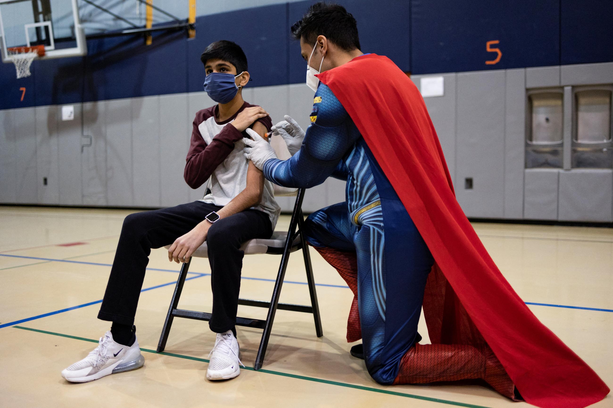 A doctor dressed in a blue Superman suit with a red cape administers a vaccine to a middle school student in a school gym