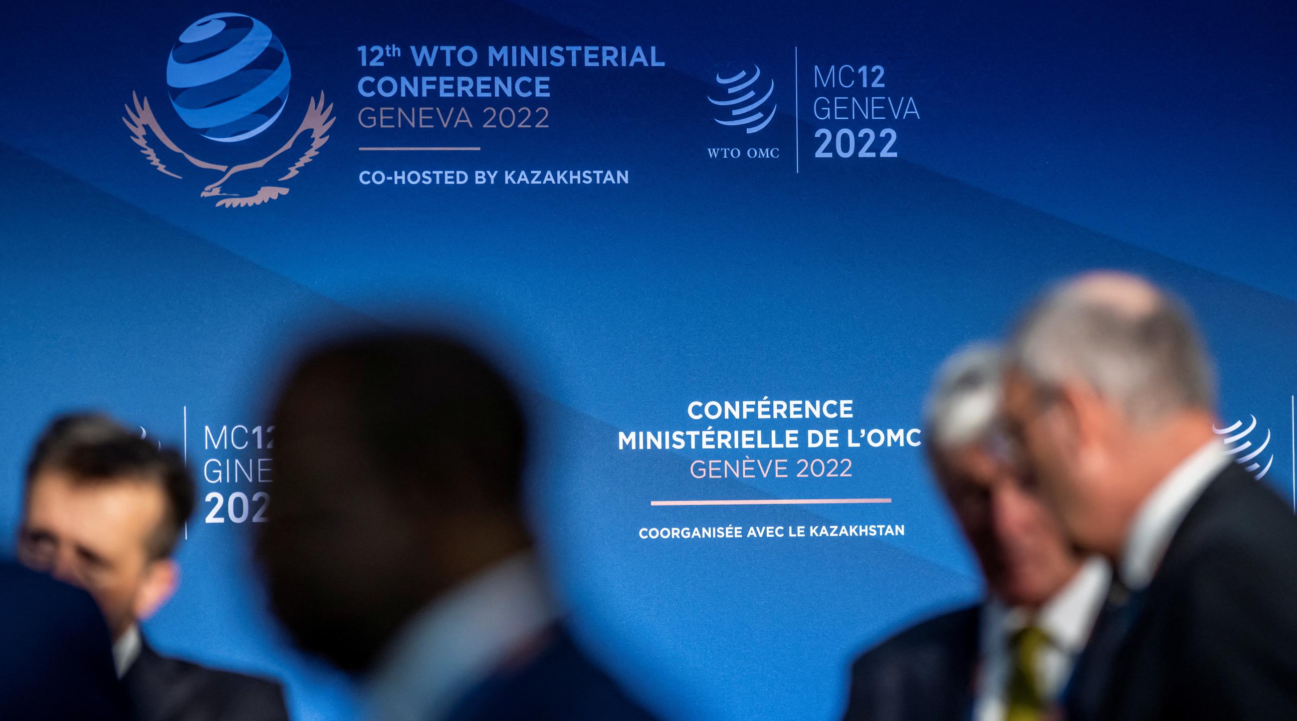 Ambassadors stand in front of a sign for the 12th Ministerial Conference at the headquarters of the World Trade Organization.