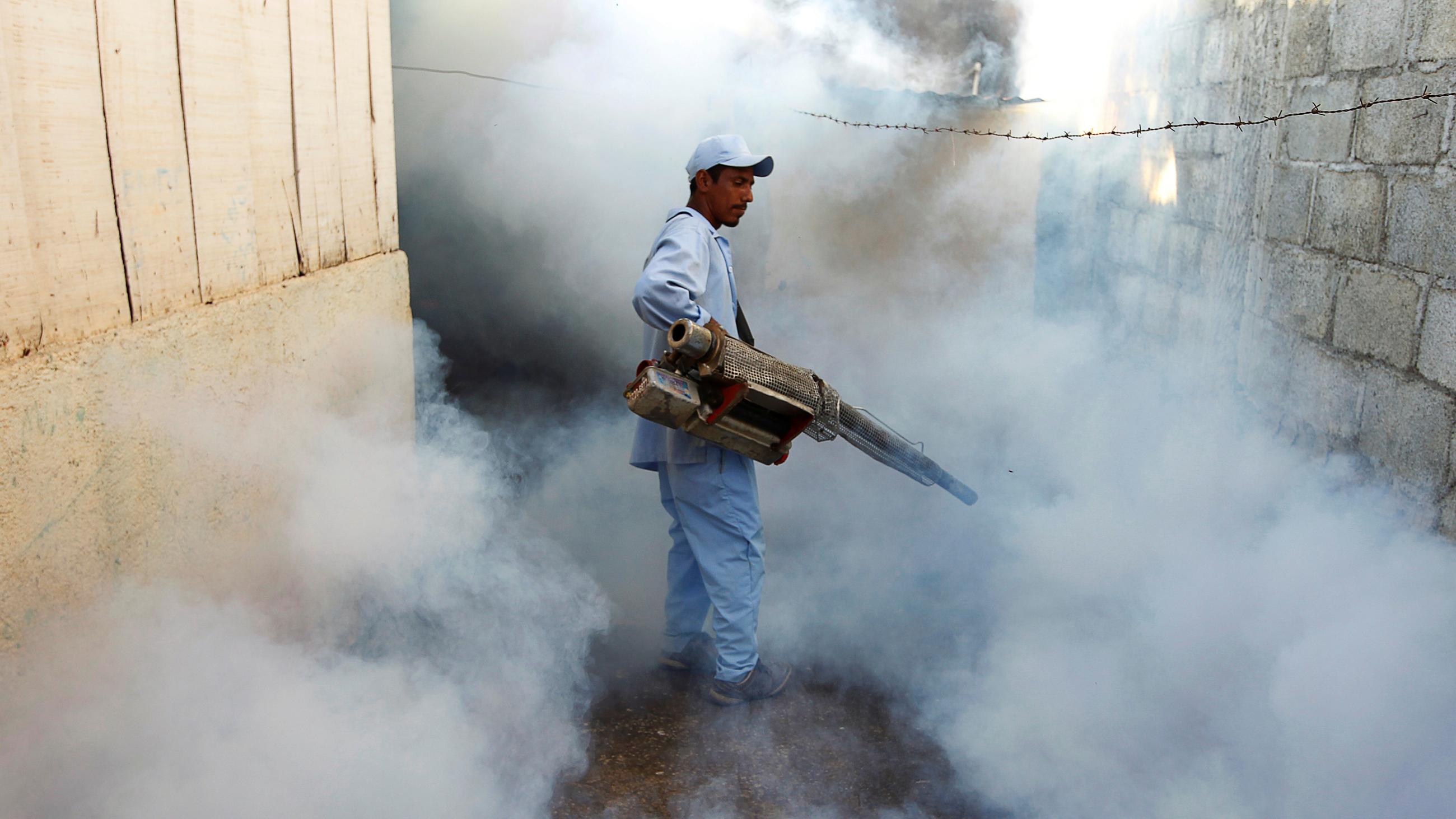 A man not wearing personal protective equipment stands in a cloud of white fumes next to a building. 