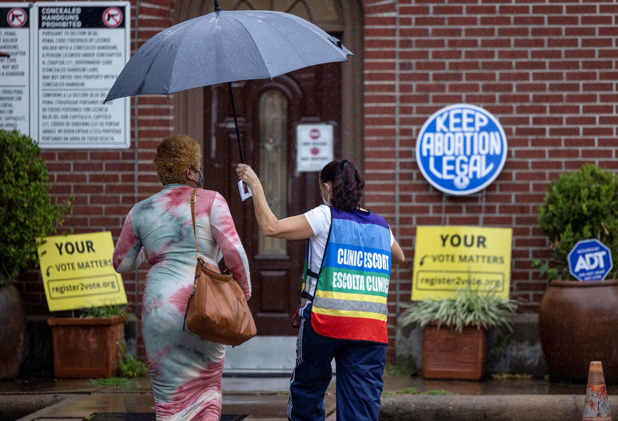 A clinic escort in a rainbow vest labeled in English and Spanish holds a black umbrella for a woman in a nice dress holding a handbag as they walk inside a red brick clinic. Outside the clinic is a blue plaque that says "Keep abortion legal" and yellow signs instructing people how to vote