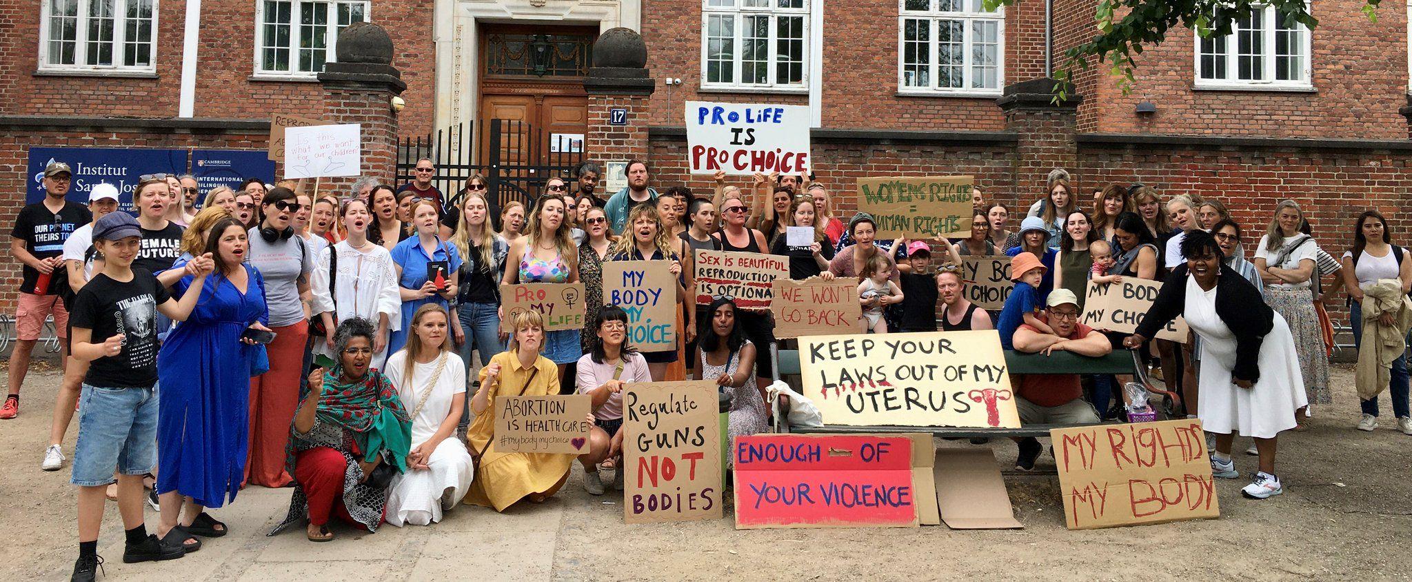 A crowd of primarily women with some men and children in summer clothing stand in front of an old brick building with cardboard signs with slogans in support of women's rights and reproductive freedom