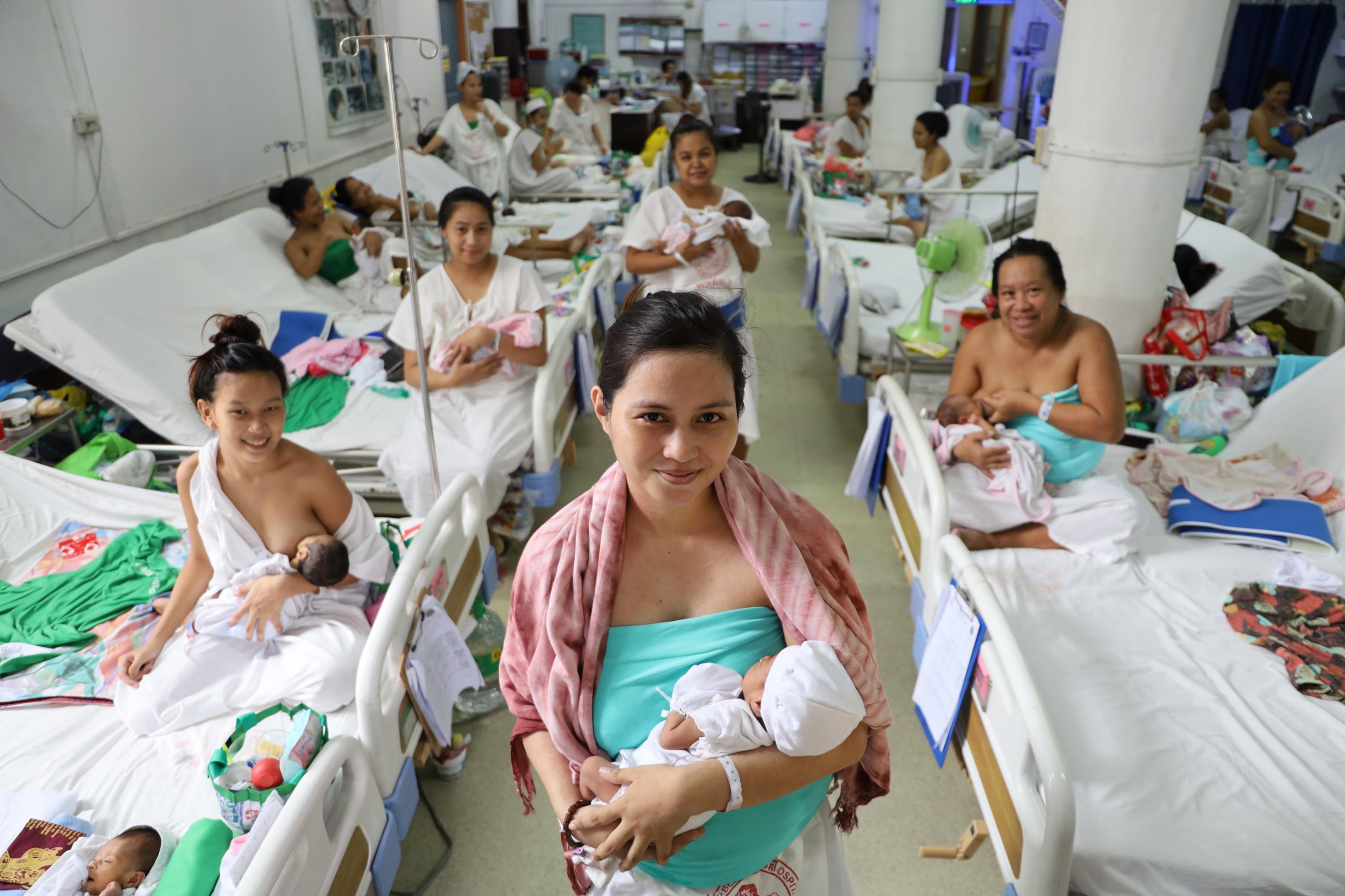 A woman in a turquoise shirt and pink shawl smiles in the center of the photograph holding her newborn infant. On either side of her are rows of hospital beds, upon which sit other women nursing infants in a maternity ward in the Philippines.