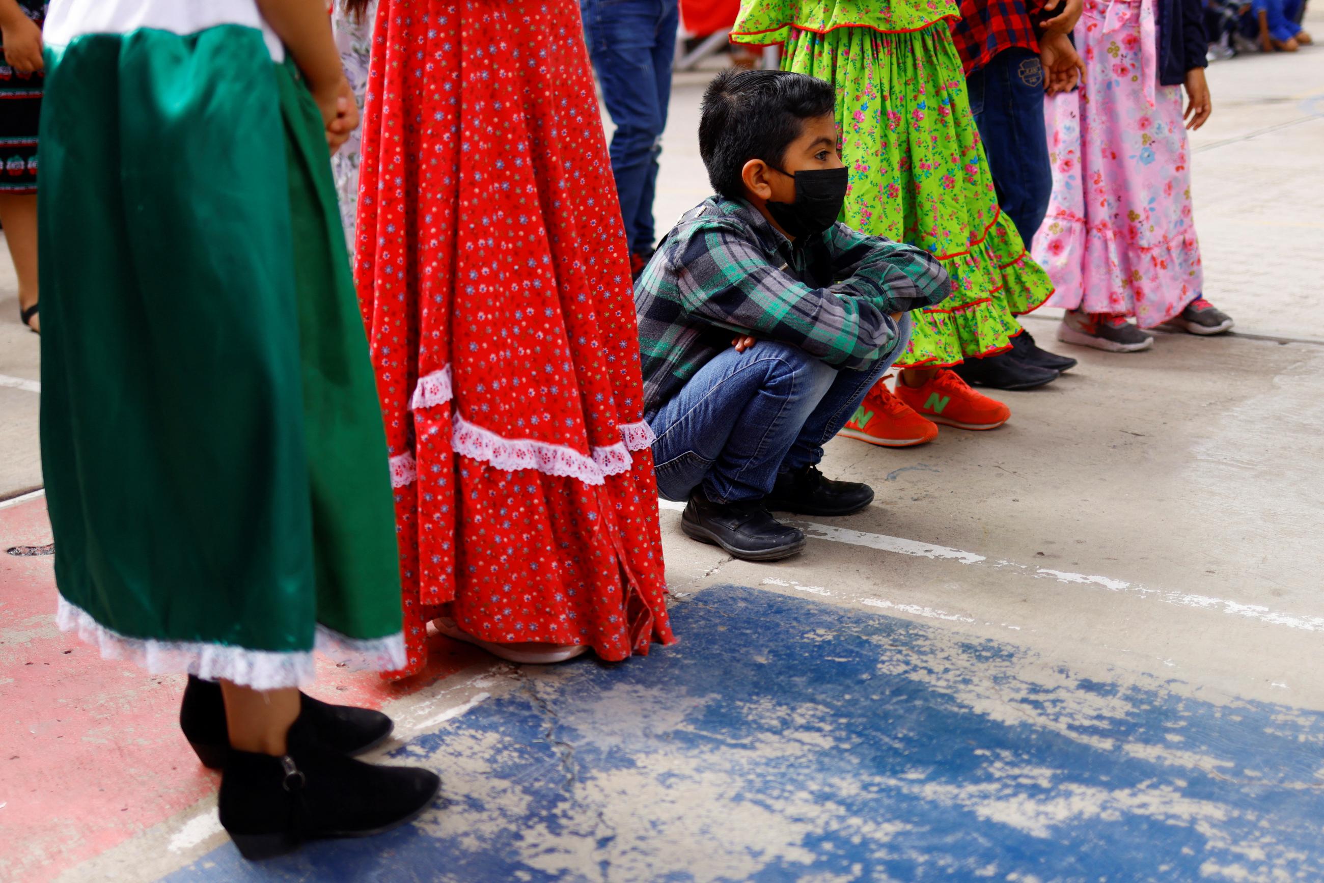A student wearing a mask squats down to rest while attending an in-person class during a COVID-19 outbreak in Ciudad Juarez, Mexico, on June 21, 2022. Colorful skirts worn by girls nearby can be seen around him.