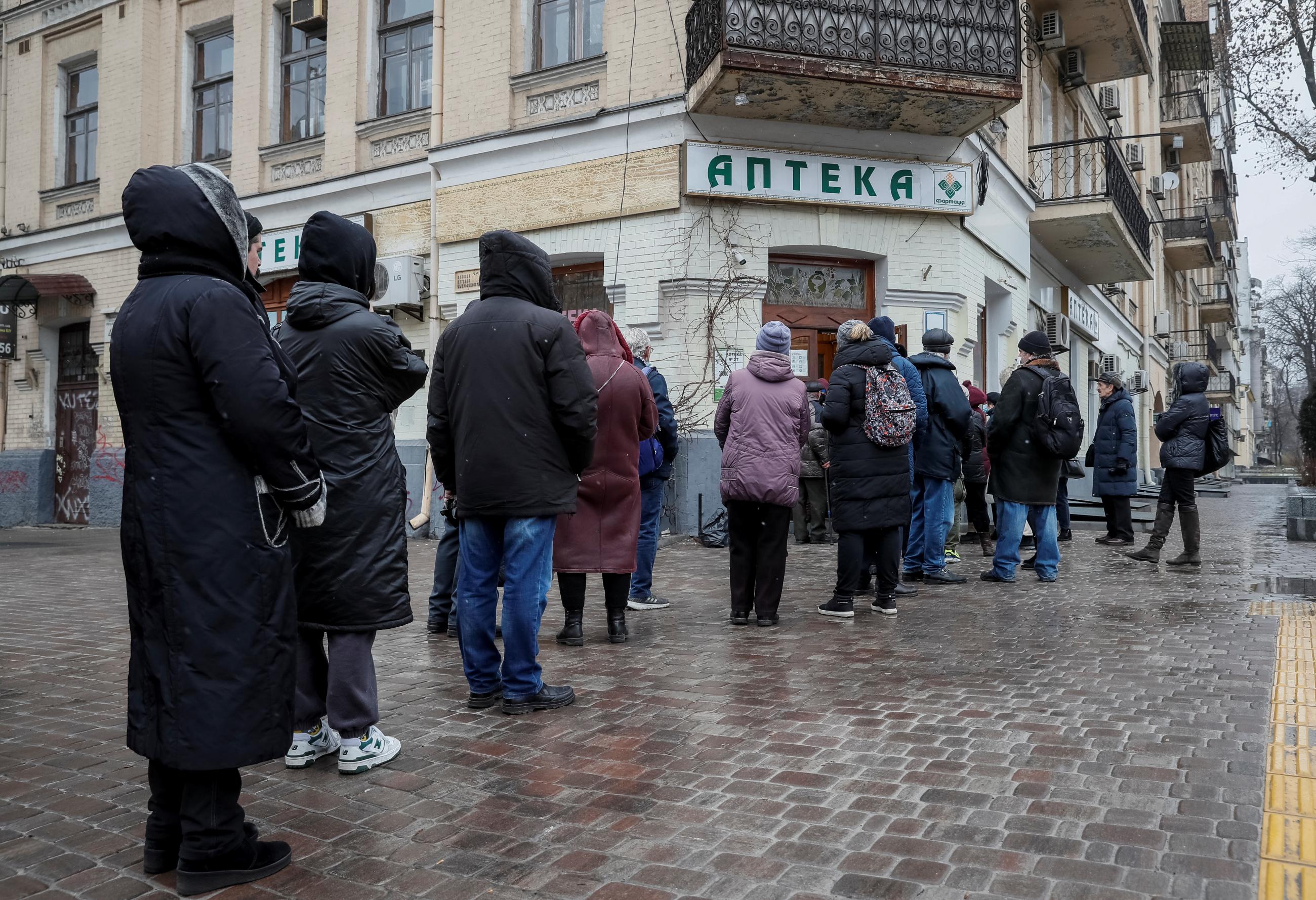 People in dark-colored winter coats line up in front of a pharmacy on a rainy spring day in Kyiv, following Russia's escalation of its war in Ukraine.