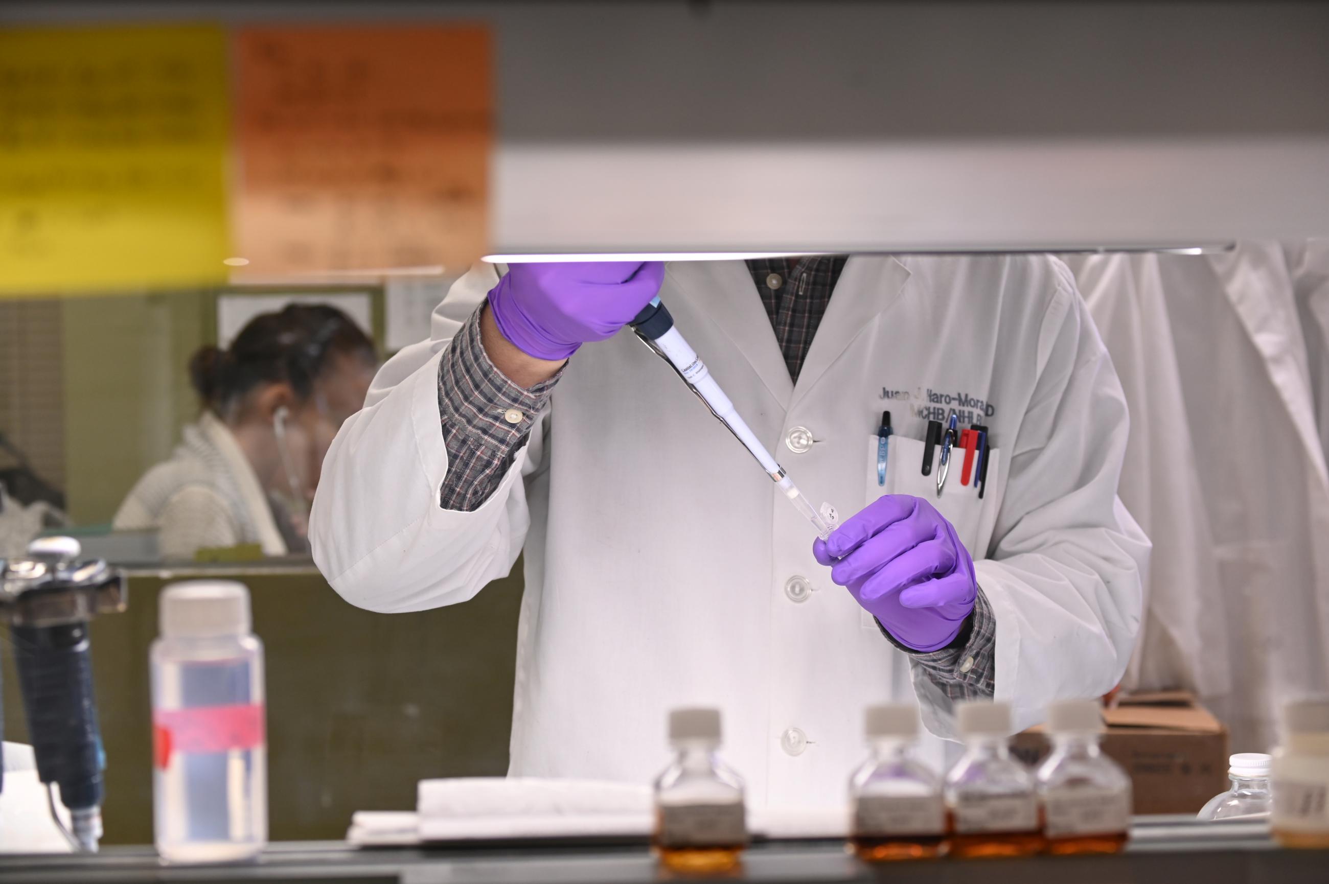 A researcher, wearing a white lab coat, whose face is obscured by white cabinetry, extracts liquid via syringe from a test tube while wearing  purple latex gloves