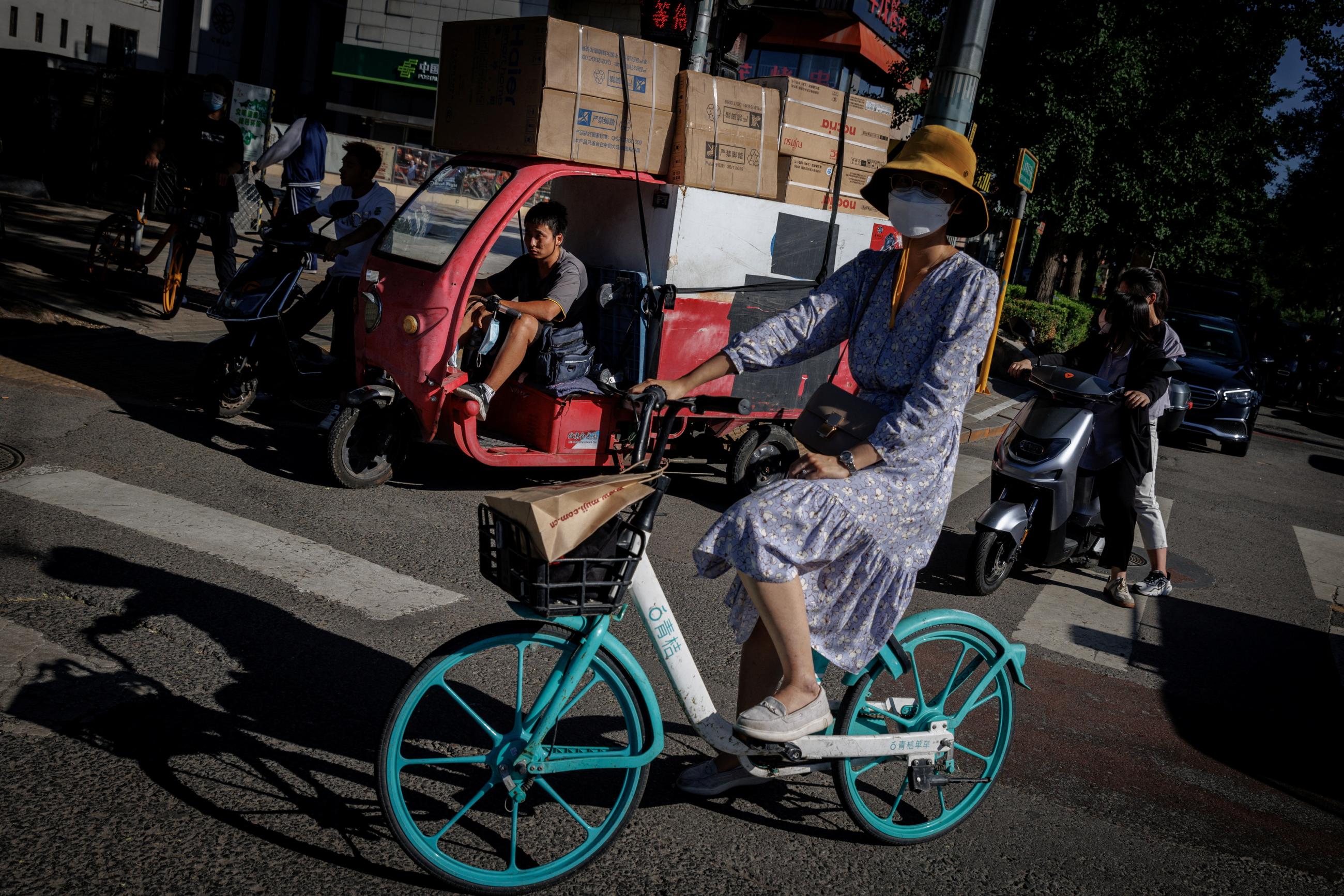 A woman on an aqua-colored bike in a dress and sneakers waits with people at a traffic light in Beijing, China, on July 14, 2022. 