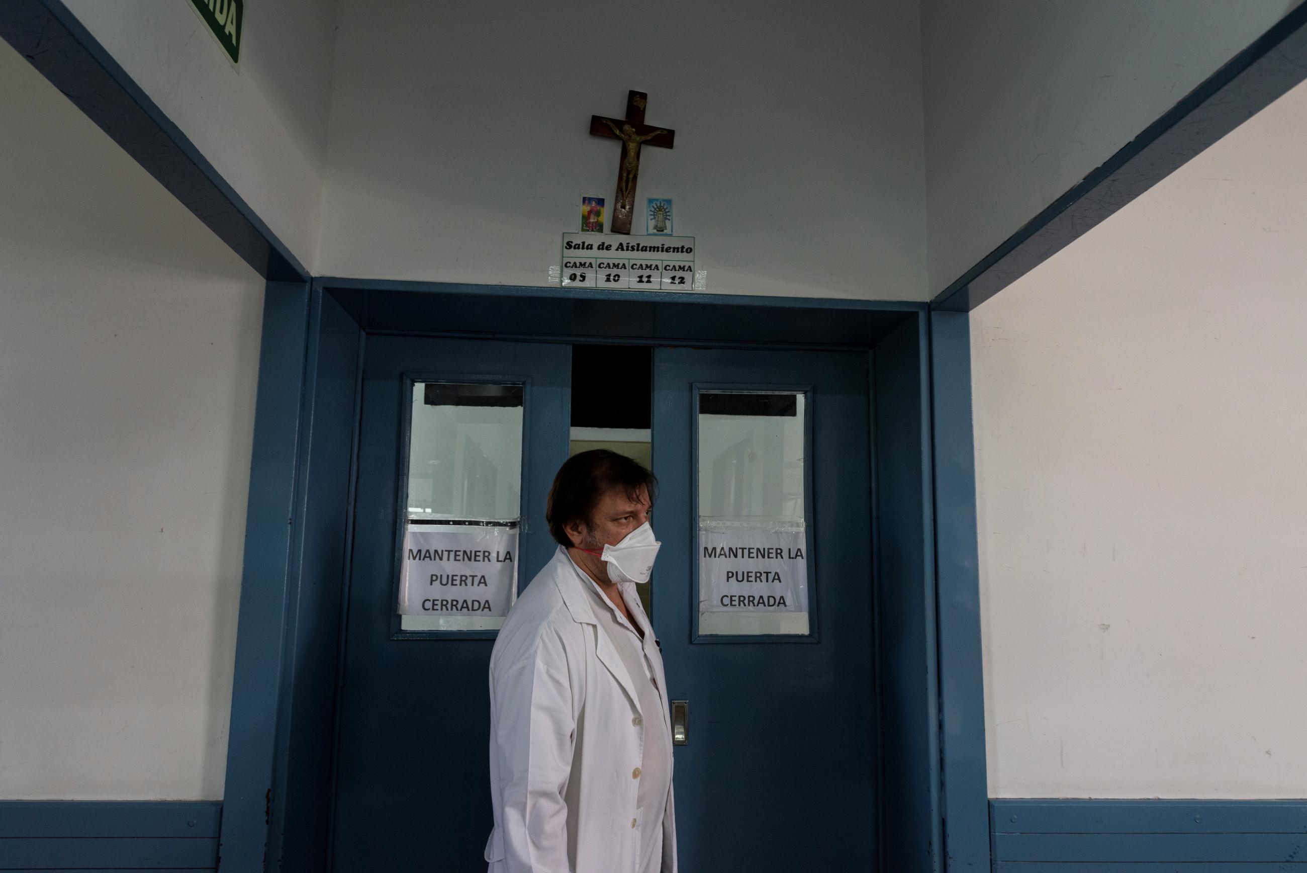Physician Luis Alberto De Carolis, an adult man with dark hair wearing a white lab coat and surgical face mask is photographed standing between two dark teal hospital doors.