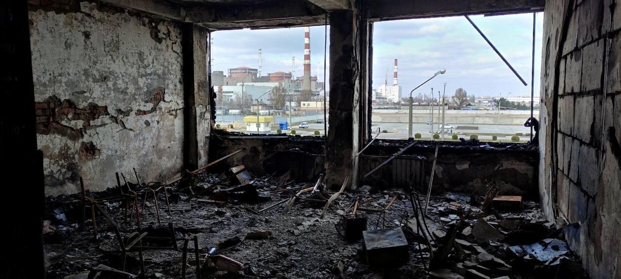 The interior of a damaged building at the Zaporizhzhia nuclear power plant compound is littered with rubble. Through the blown-out windows can be seen the red and white towers of the nuclear facility.