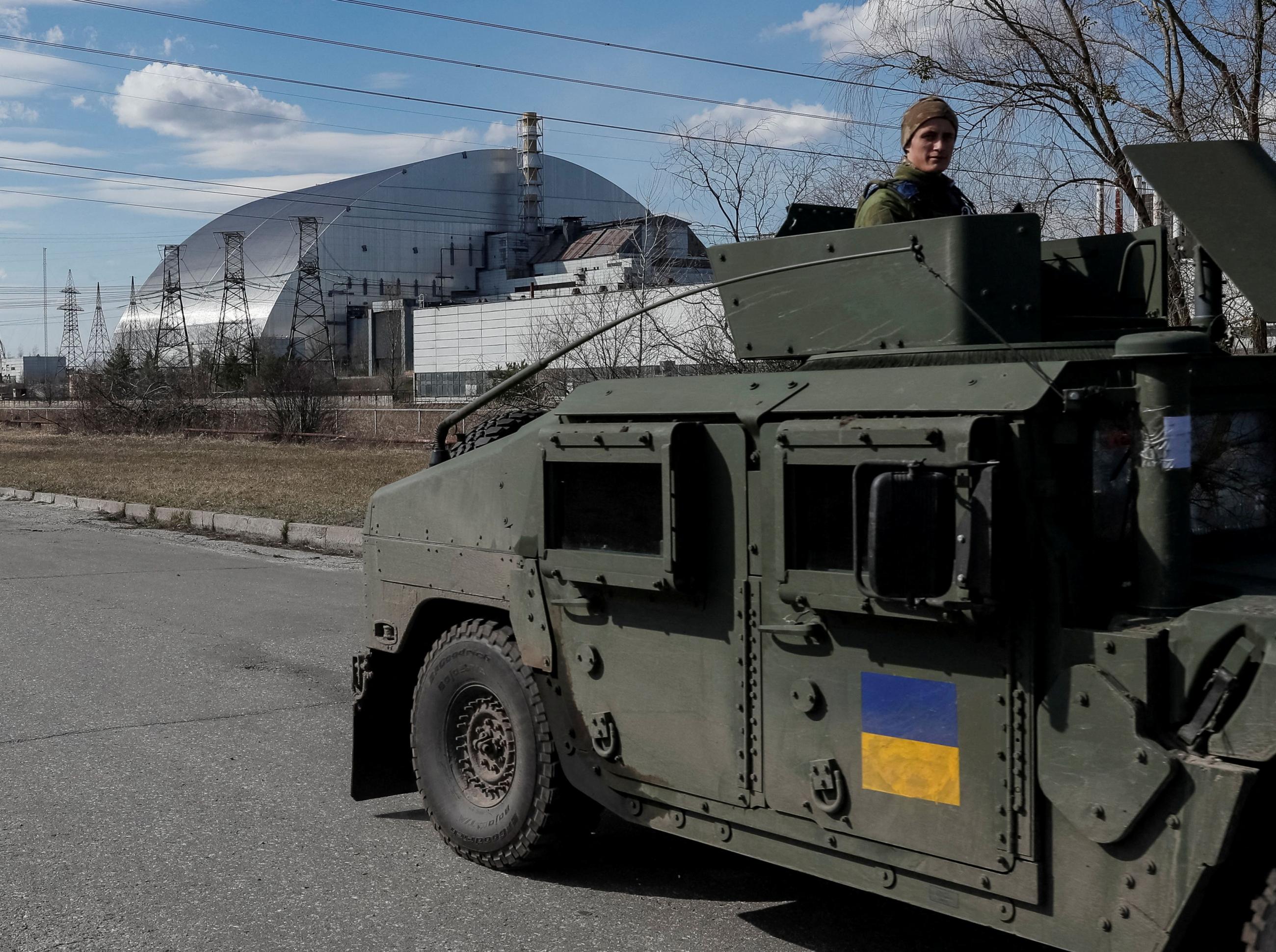 Ukrainian national guardsmen drive a green military vehicle emblazoned with the blue and yellow Ukrainian flag patrol the area near the Chernobyl Nuclear Power Plant. In the background looms the silver dome of a nuclear power plant building. 
