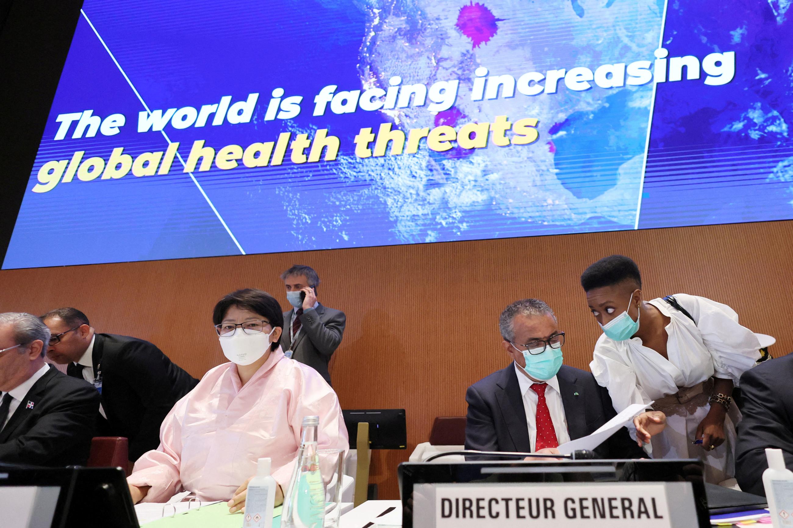 Dr. Tedros Adhanom Ghebreyesus is photographed sitting behind a podium wearing a dark suit, white shirt, and red tie flipping through a newspaper at the 75th World health Assembly meeting
