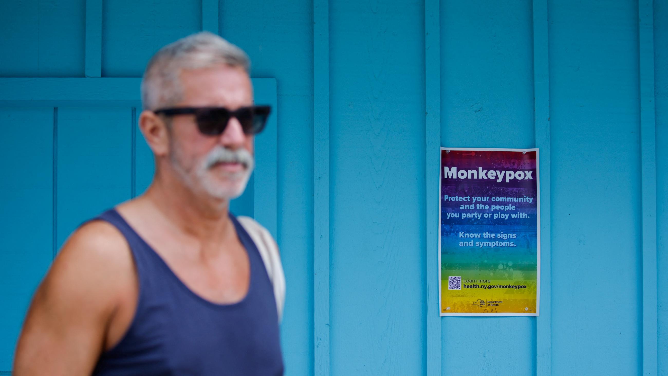 A grey-haired bearded man in a blue tank top and sunglasses walks past a turquoise wall with a rainbow poster that reads "Protect your community and the people you party or play with. Know the signs and symptoms" placed there by the New York Department of Health