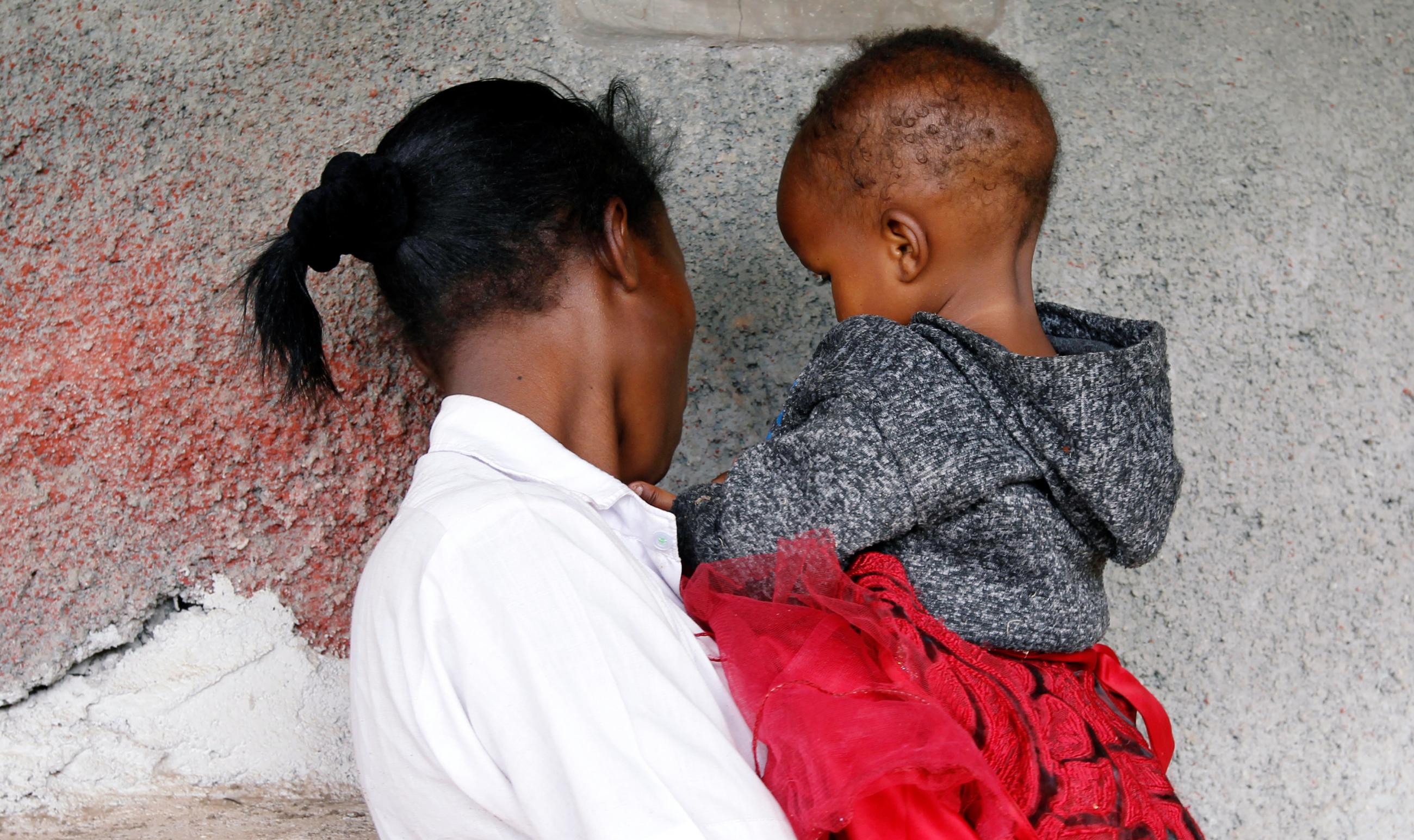 A young woman wearing a white shirt is photographed from behind holding her baby weraing a gray jumper