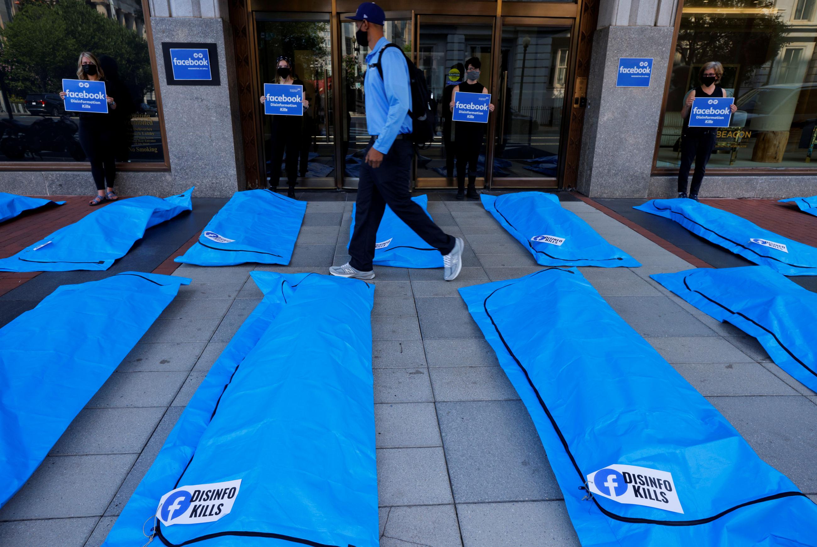 A man in a blue shirt and black mask walks through an art installation of bright blue body bags laid out on the sidewalk with labels that say "Disinfo kills"