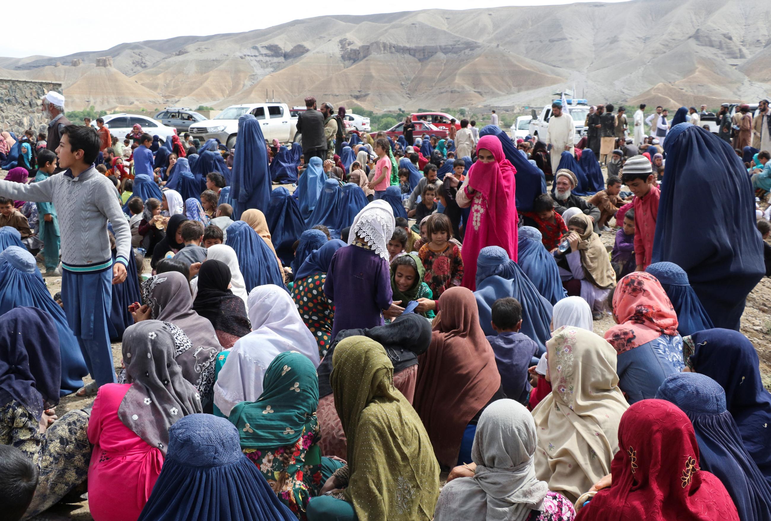 A crowd of displaced Afghan families in blue and pink clothing spend time together after the heavy flood in the Khushi district of Logar, Afghanistan. In the background, mountains rise up from the horizon.