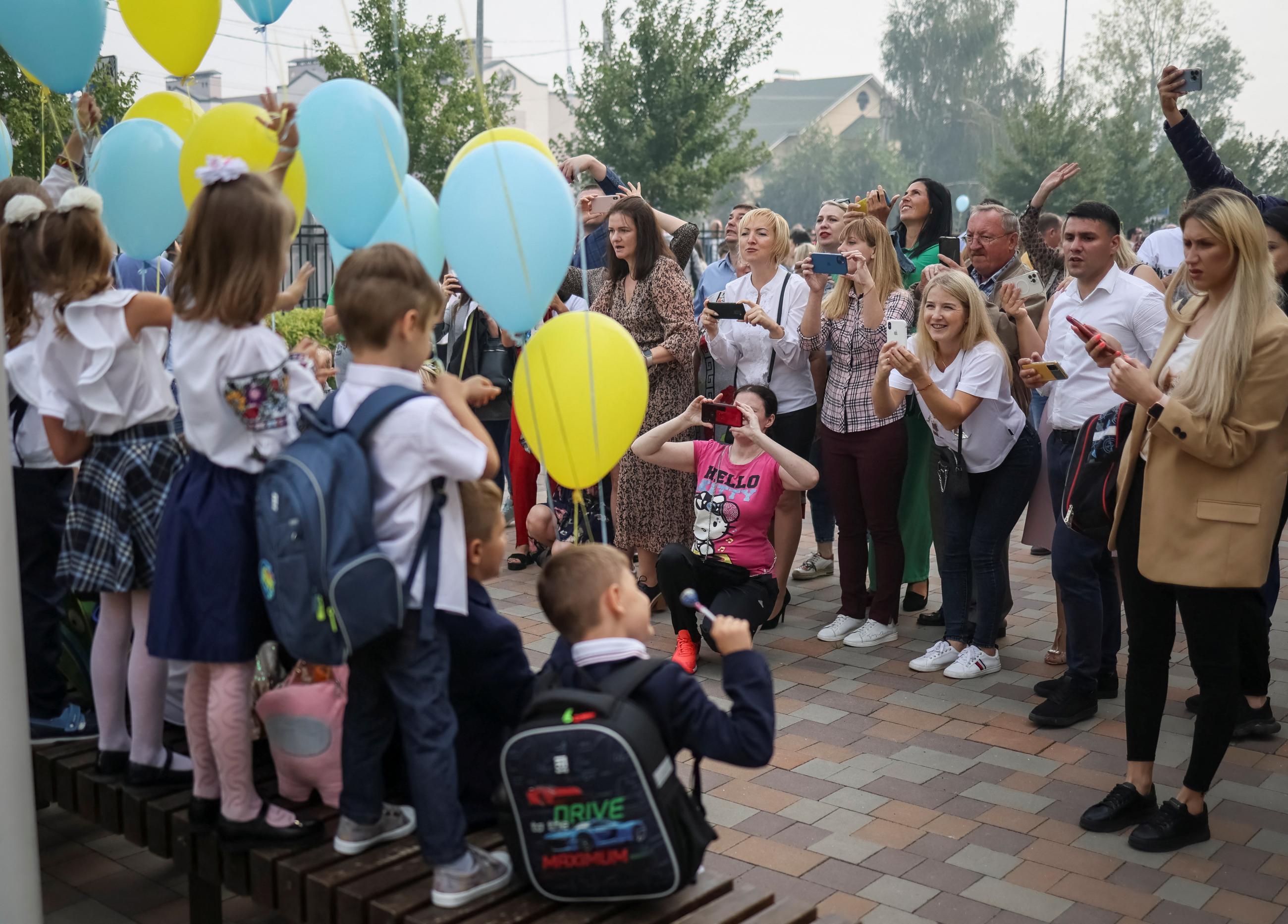 Parents with smartphones take pictures of their elementary-school-aged children, who are wearing school uniforms and backpacks and holding blue and yellow balloons during a ceremony to mark the start of the school year. The children have their backs to the camera, but are posing happily for their smiling parents.