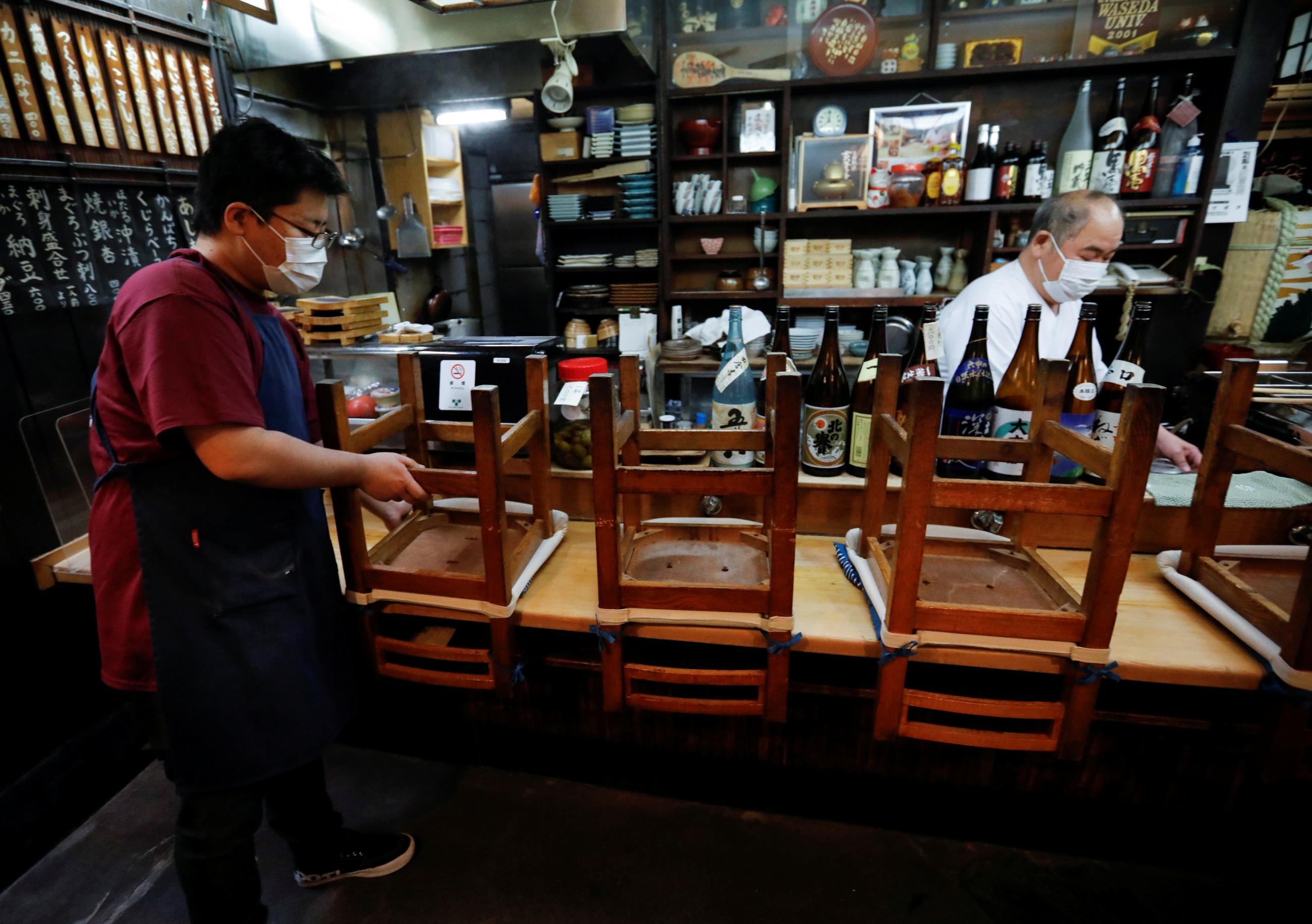 A young man in a red t-shirt stacks chairs while an older man cleans up the bar area of an Izakaya, a Japanese-style dining bar. In the background and on the bar are displayed tall bottles of sake with colorful labels