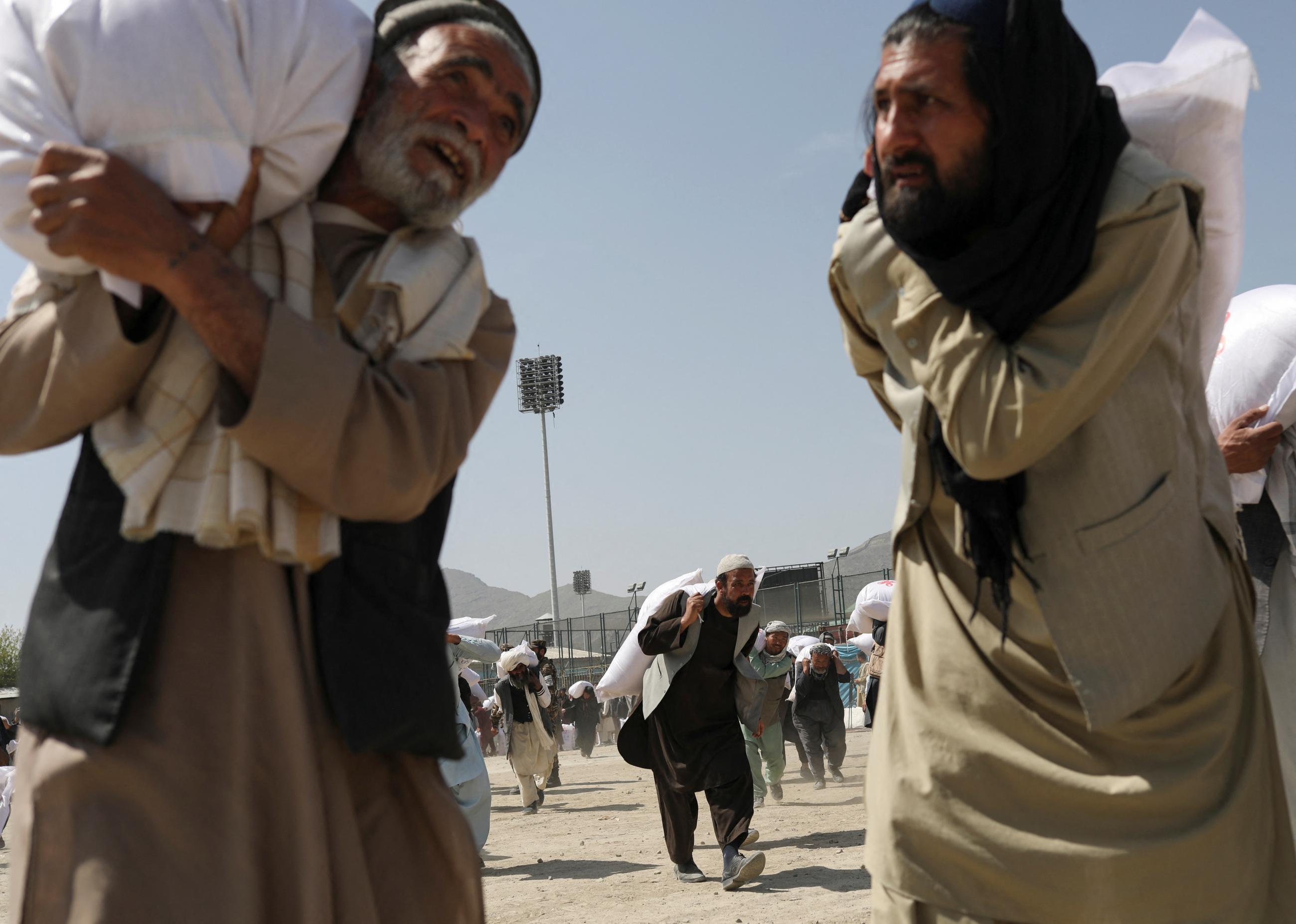 In the foreground of the photo, two men with beards carry large sacks of rice over their shoulders, while other men carry sacks in the background. The rice donations are part of a humanitarian aid effort, sent by China to Afghanistan, at a distribution center in Kabul, Afghanistan, on April 7, 2022.