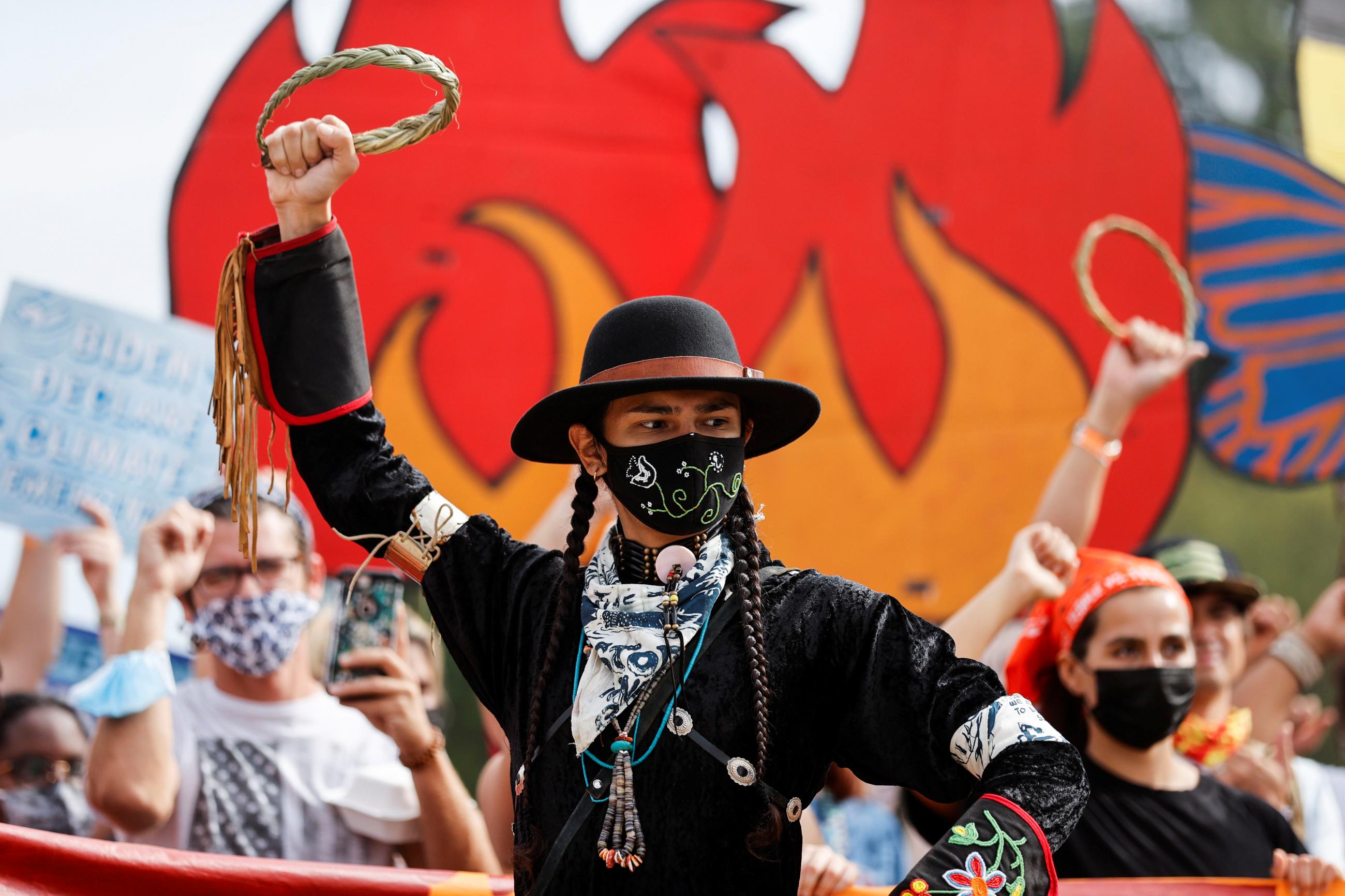 A giant poster of a red and orange flame is seen behind an environmental activist dressed in a black hat and jacket, who is lifting his arm in the air and carrying a braided corn husk, at a climate change protest near the U.S. Capitol in Washington, D.C., on October 15, 2021.