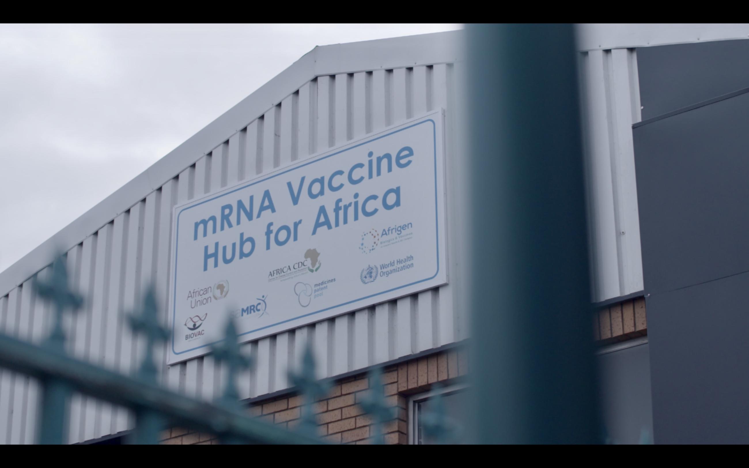 The side of an industrial looking building features a sign that says, "mRNA Vaccine Hub for Africa." On the bottom of the sign are the names of "hub" partners and their logos, including the African Union, Africa CDC, Afrigen, Biovac, MRC, and the World Health Organization.