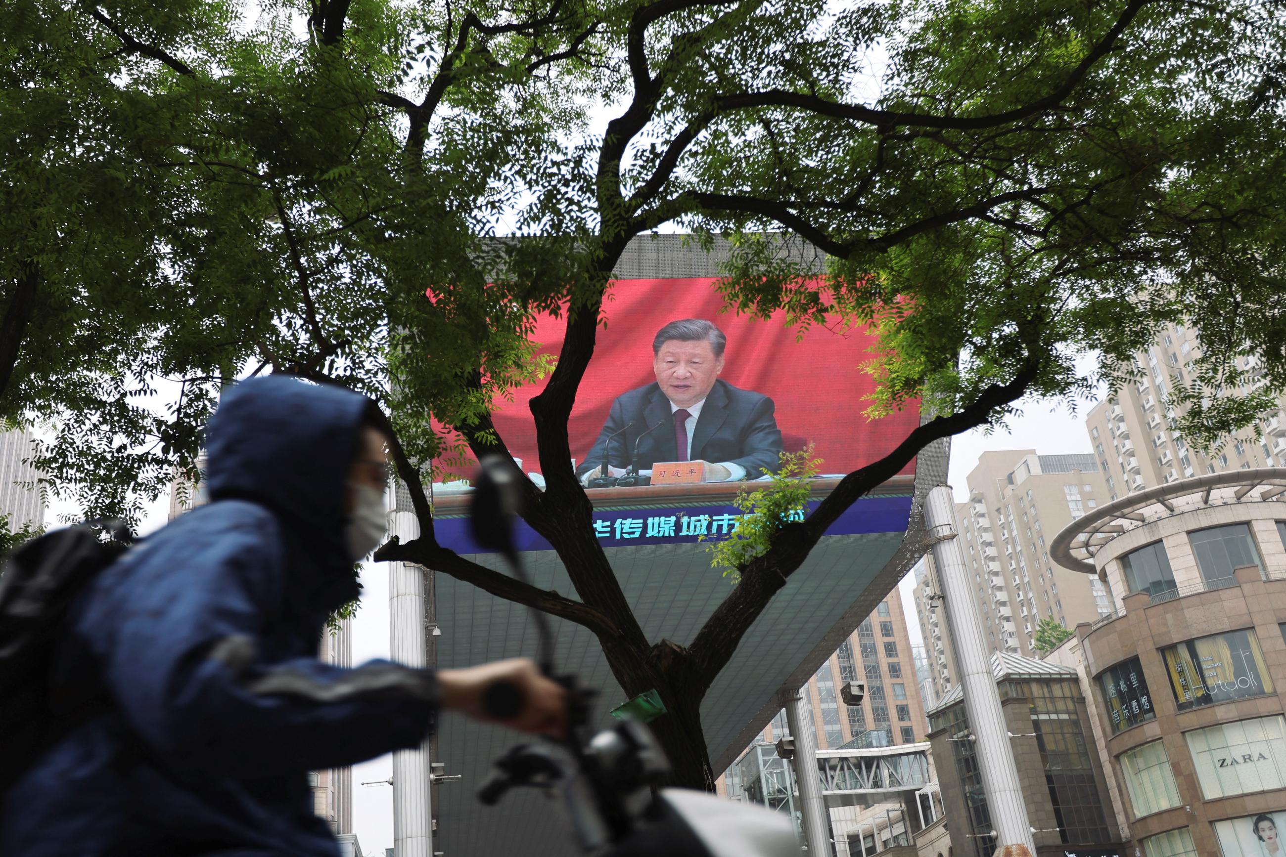 A man wearing a face mask rides past a giant screen showing Chinese President Xi Jinping speaking.