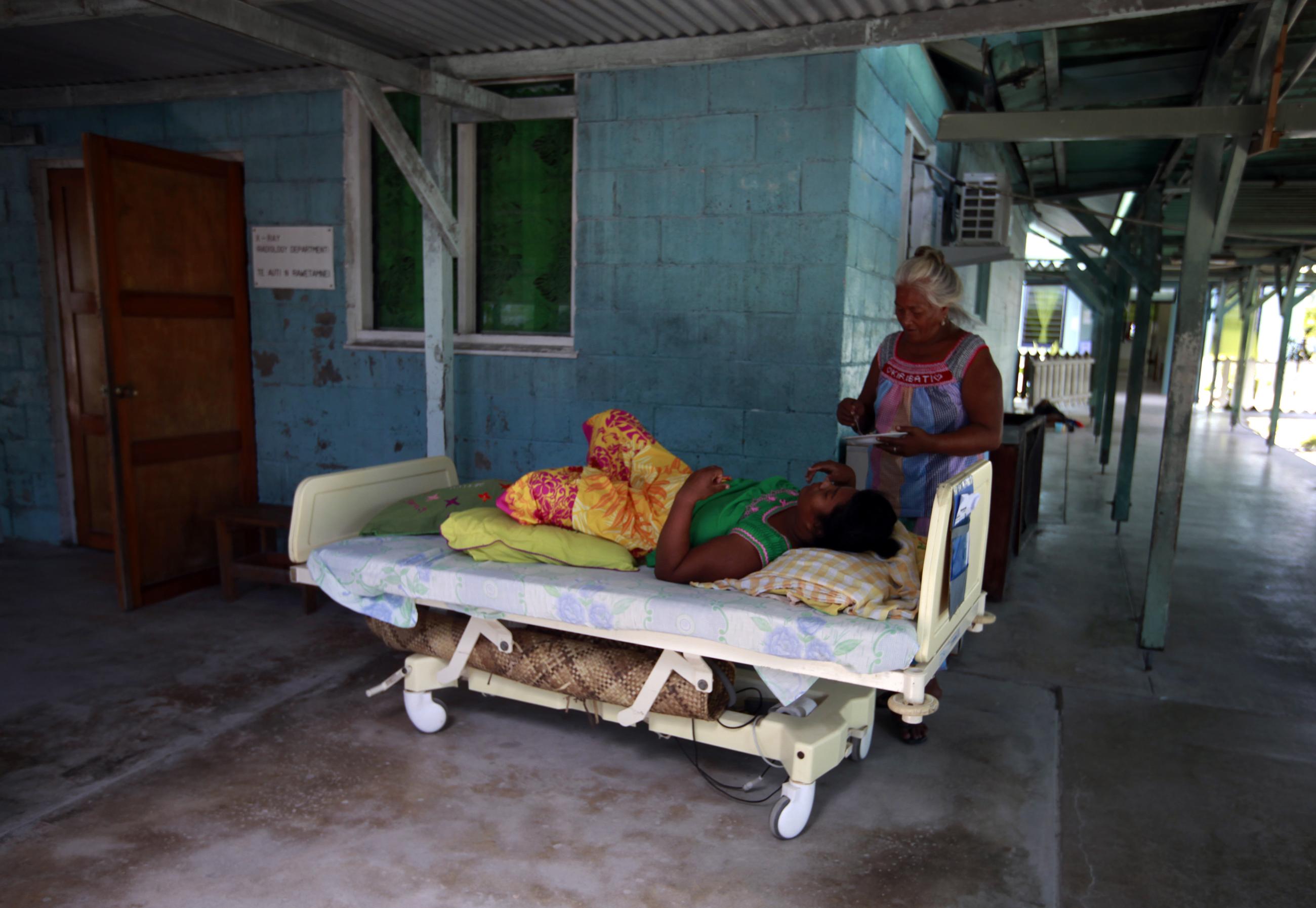 A woman prepares to feed a patient on a bed, placed outside due to lack of space.