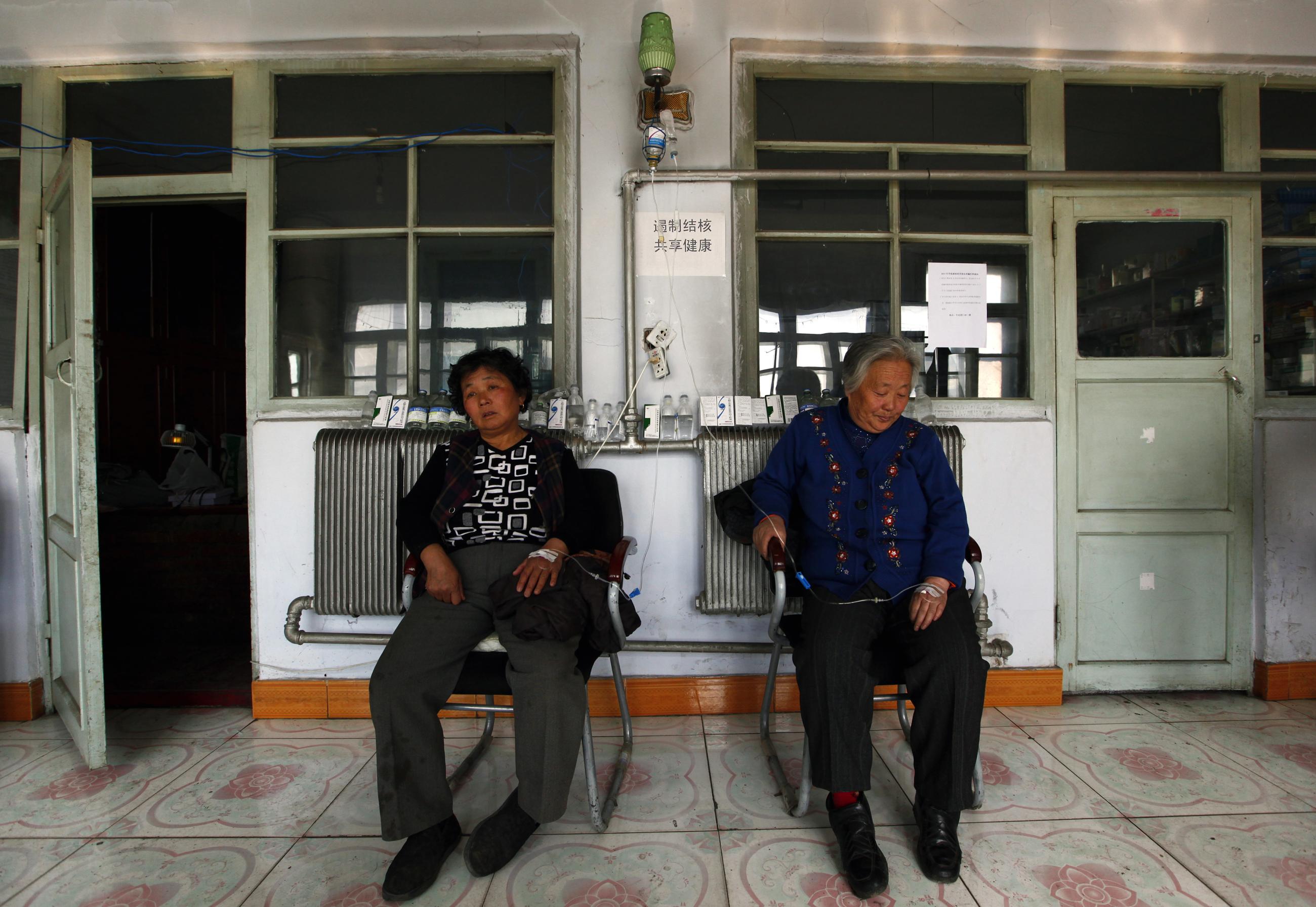 Patients sit with drips in their hands that hang from a light-fitting above them.